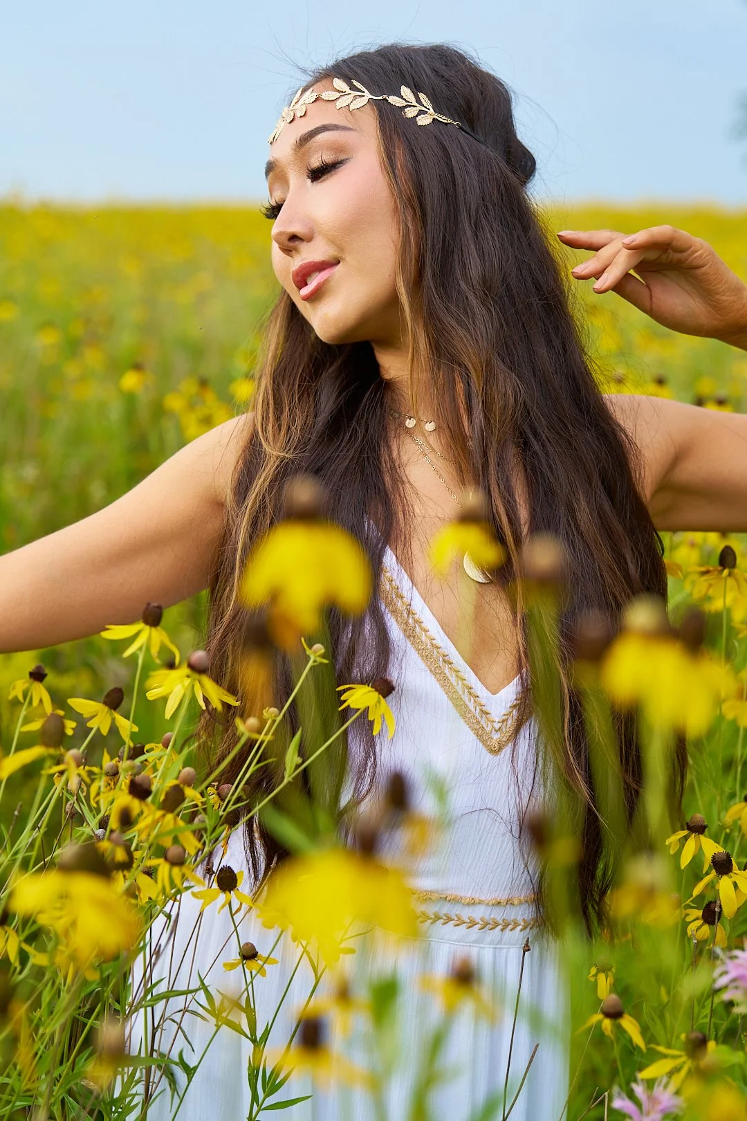 A woman with long dark hair wearing a white dress with gold embroidery, a gold leaf headband, and layered necklaces, standing in a field of yellow flowers under a clear blue sky.