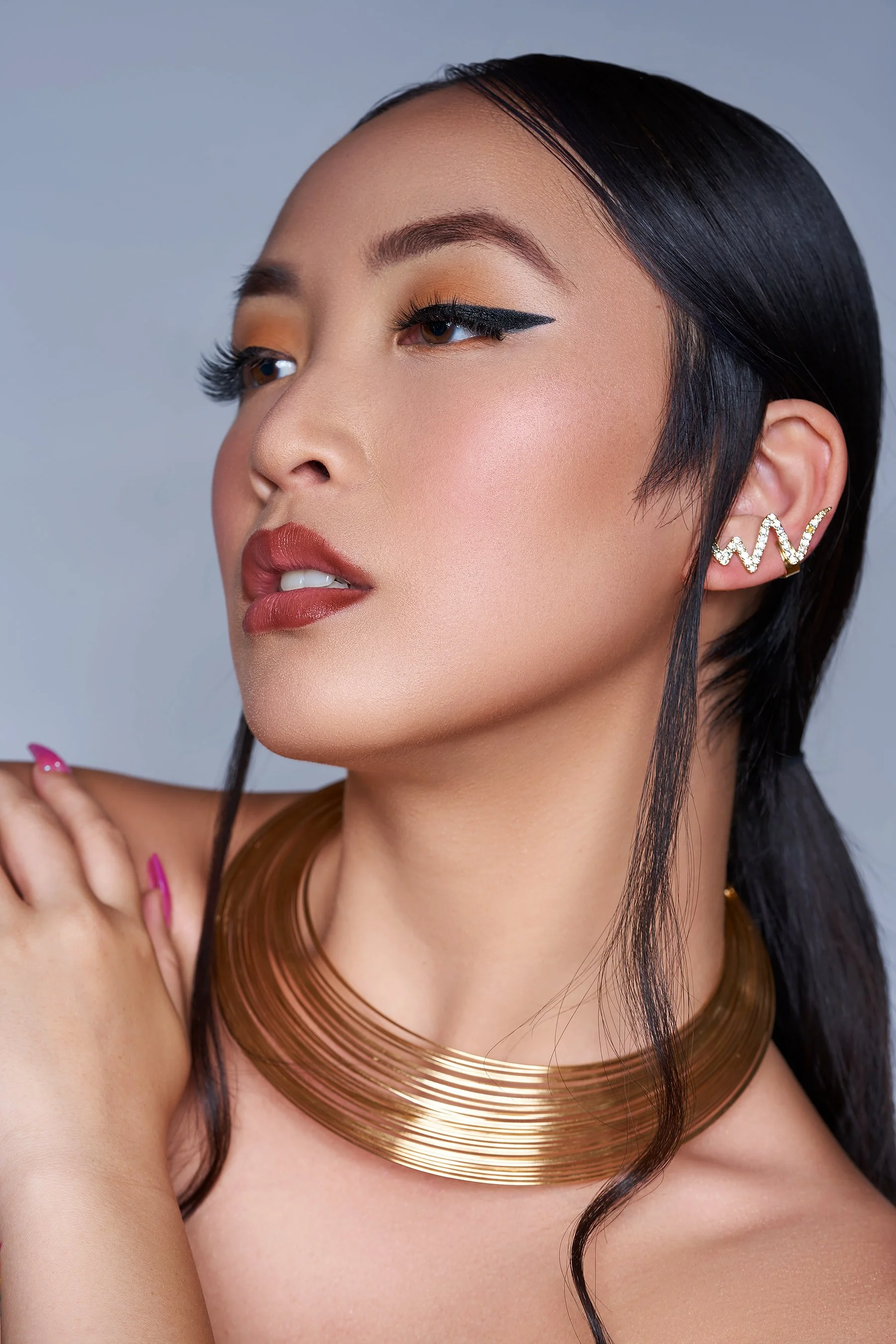 Close-up of a woman with black hair, wearing bold makeup, gold and pink jewelry, and a gold statement necklace.