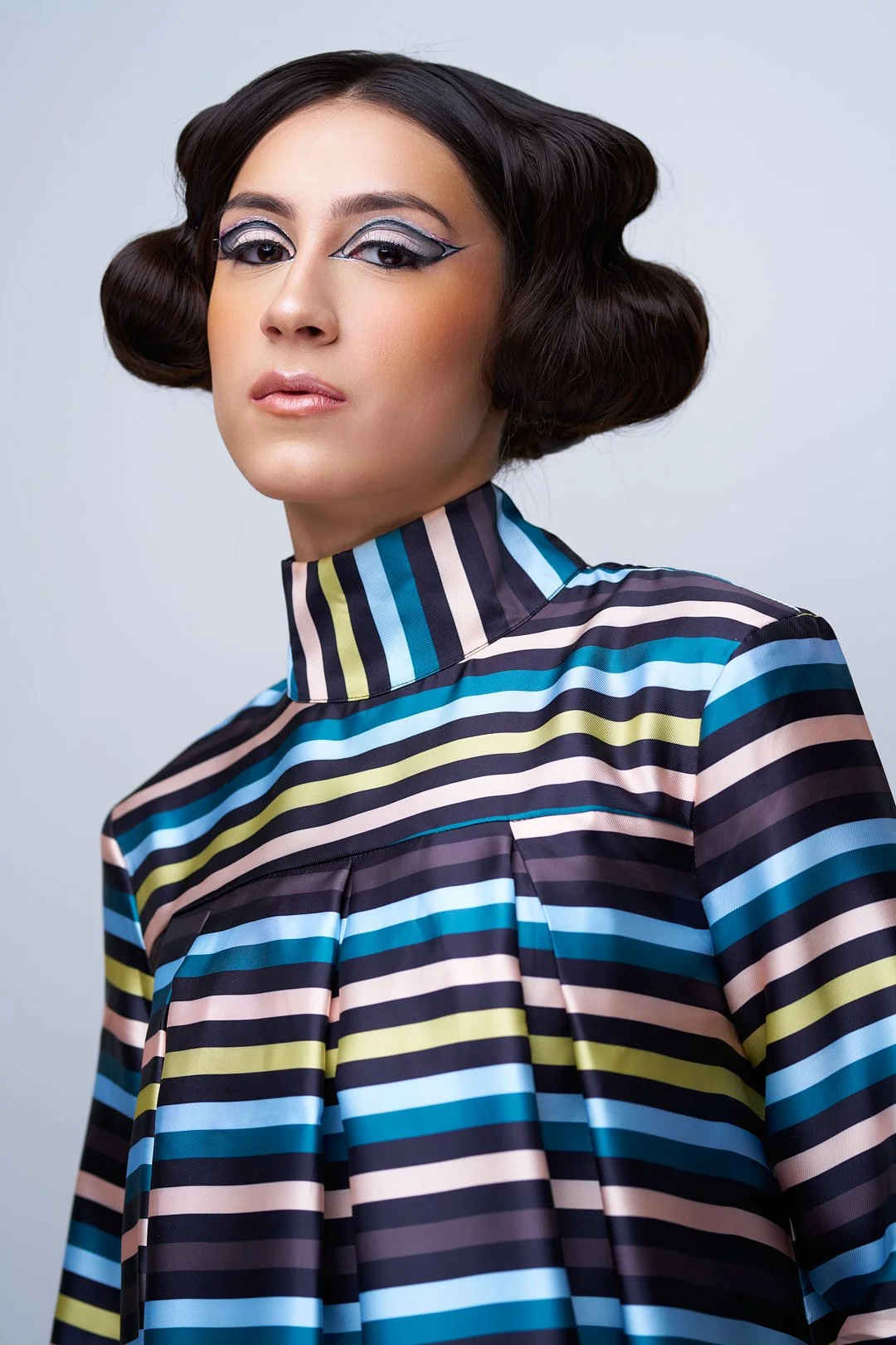 A woman with styled dark hair and artistic makeup wearing a multicolored striped high-collared dress.