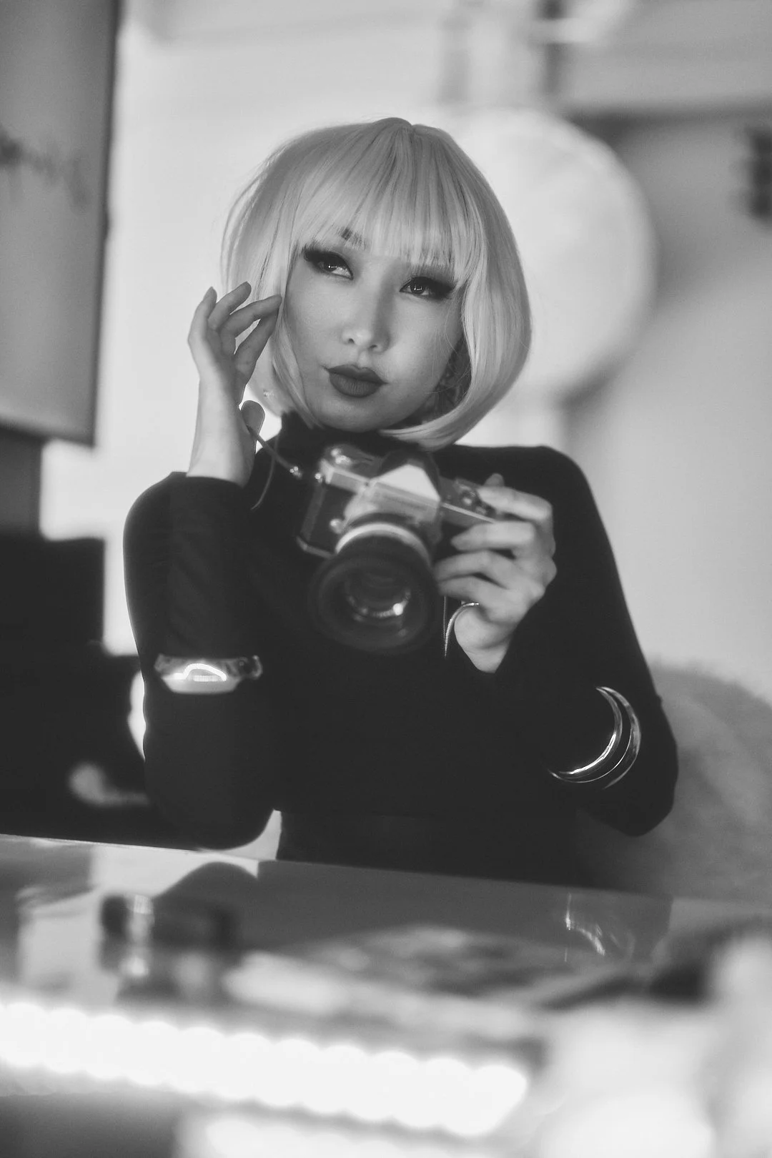 A woman with a blonde bob haircut and dark makeup holding a vintage camera, looking sideways with a contemplative expression.