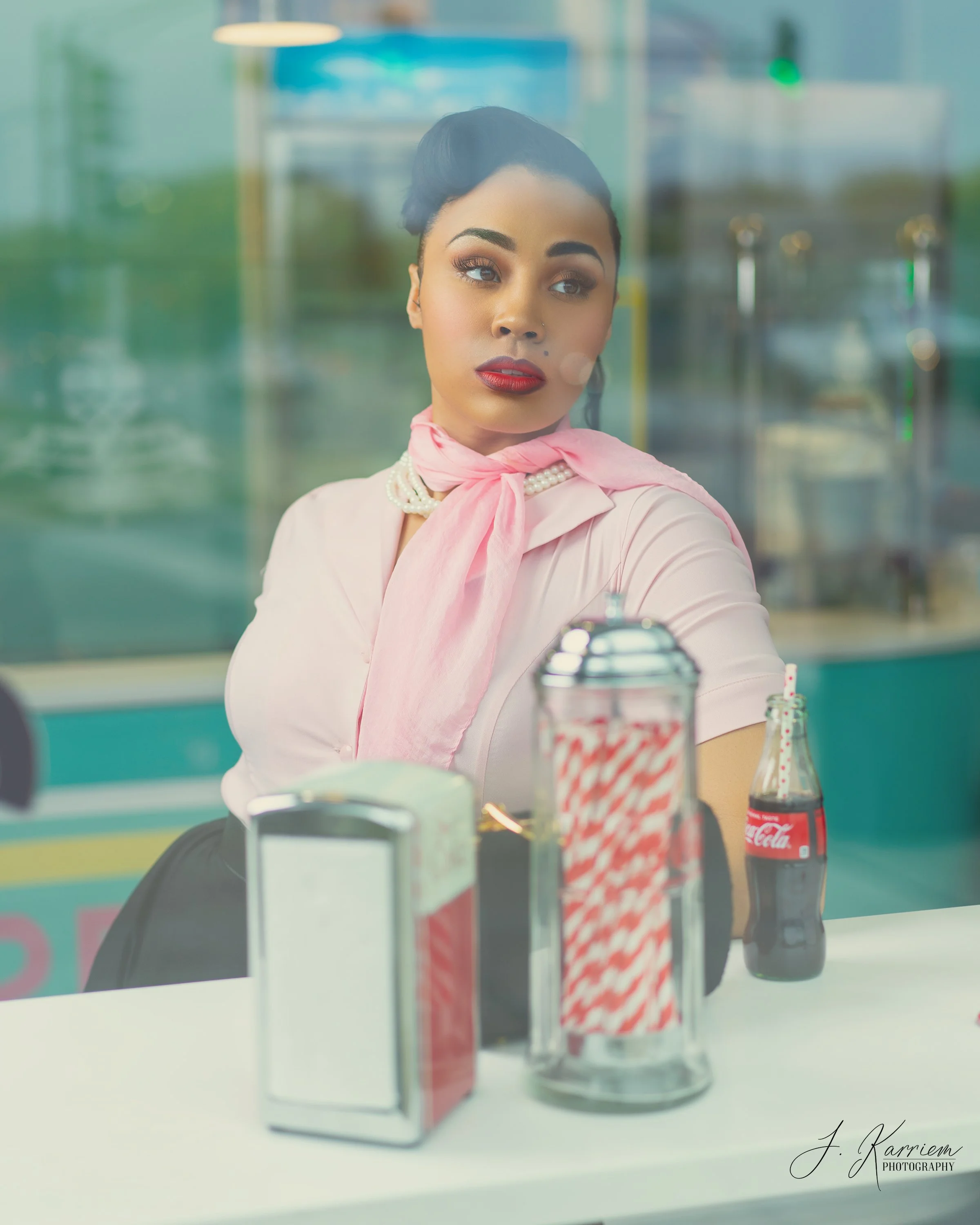 A woman with dark hair styled in an updo, wearing lipstick, pearl necklace, pink scarf, and pink blouse, looking out through a window in a retro-style diner. Visible items on the counter include a red and white straw dispenser, a glass soda bottle, a