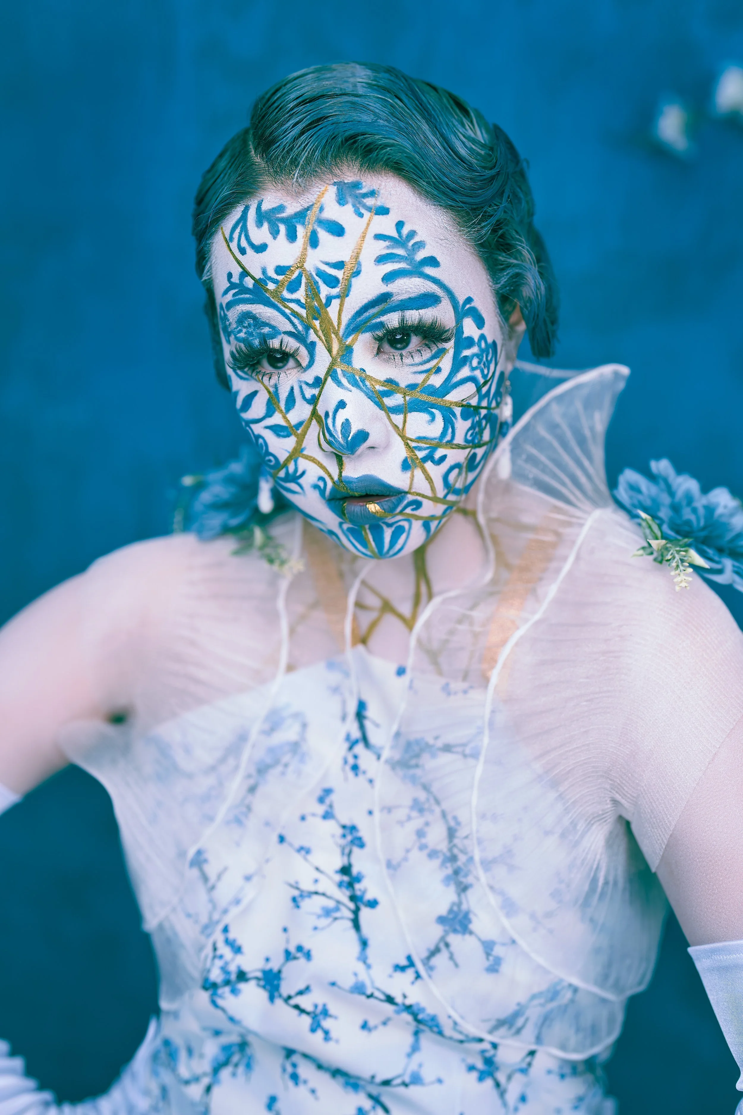 A person with elaborate blue and gold face paint, wearing a sheer white garment with floral designs, set against a blue background.