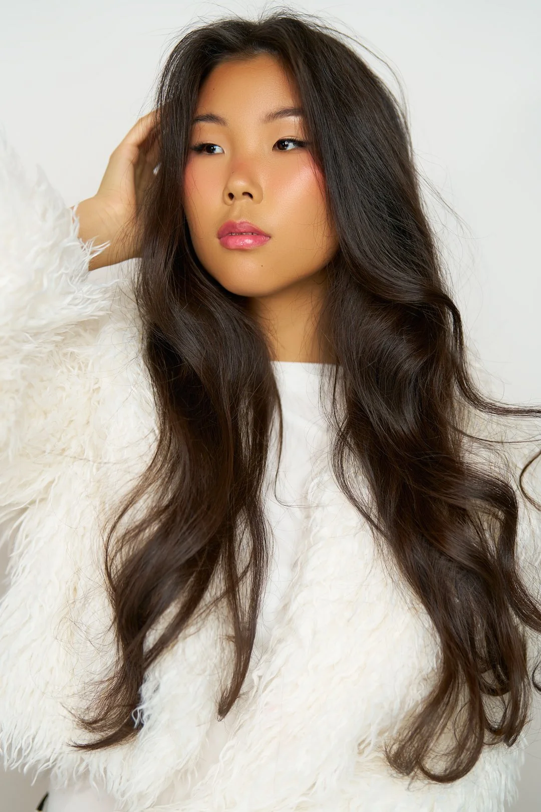 A young woman with long, wavy dark hair, wearing a white, fluffy top, posing with one hand near her head against a plain background.