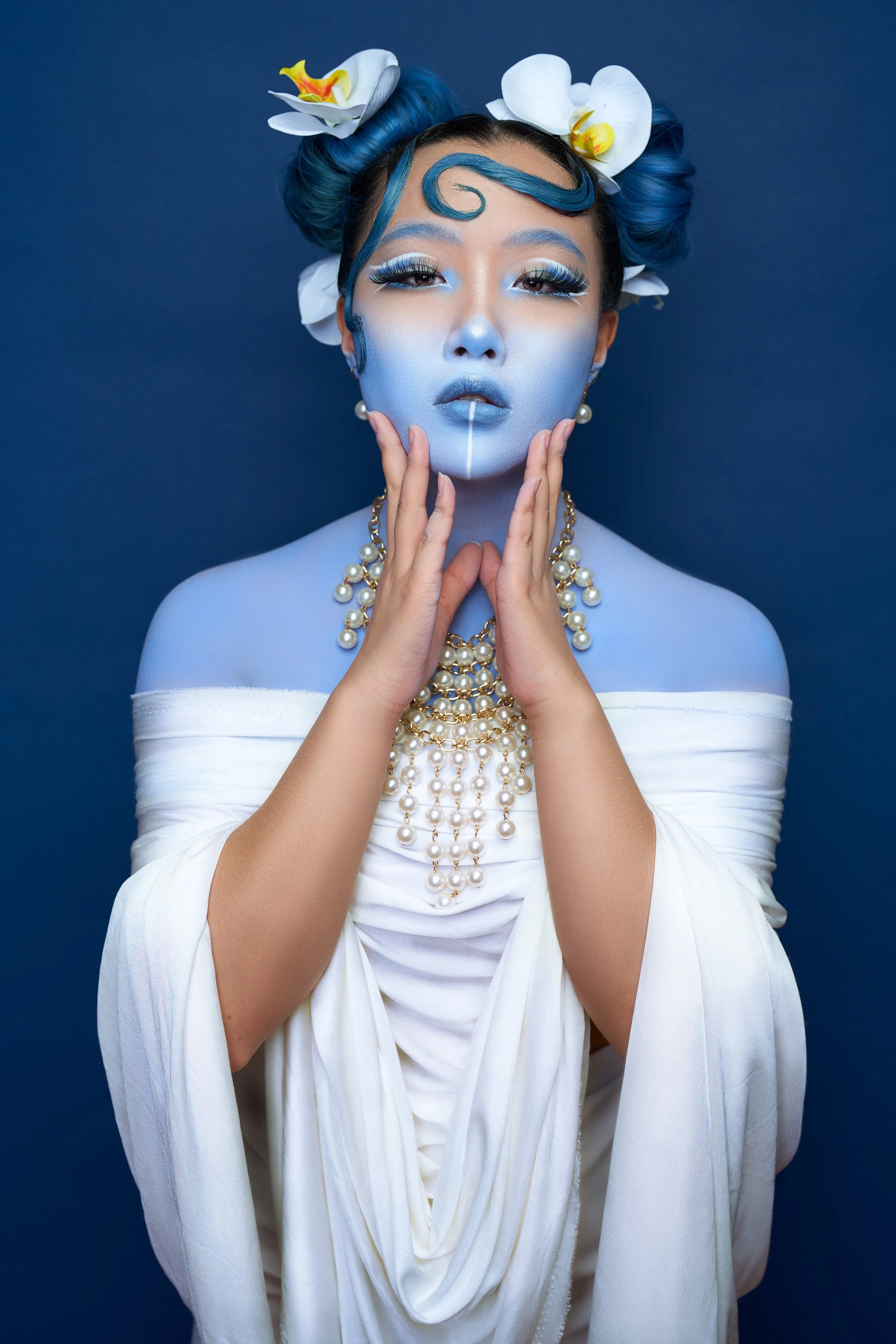 Woman with blue skin makeup and blue hair styled in buns with white orchids, wearing pearl jewelry, holding her face with both hands, against a solid dark blue background.