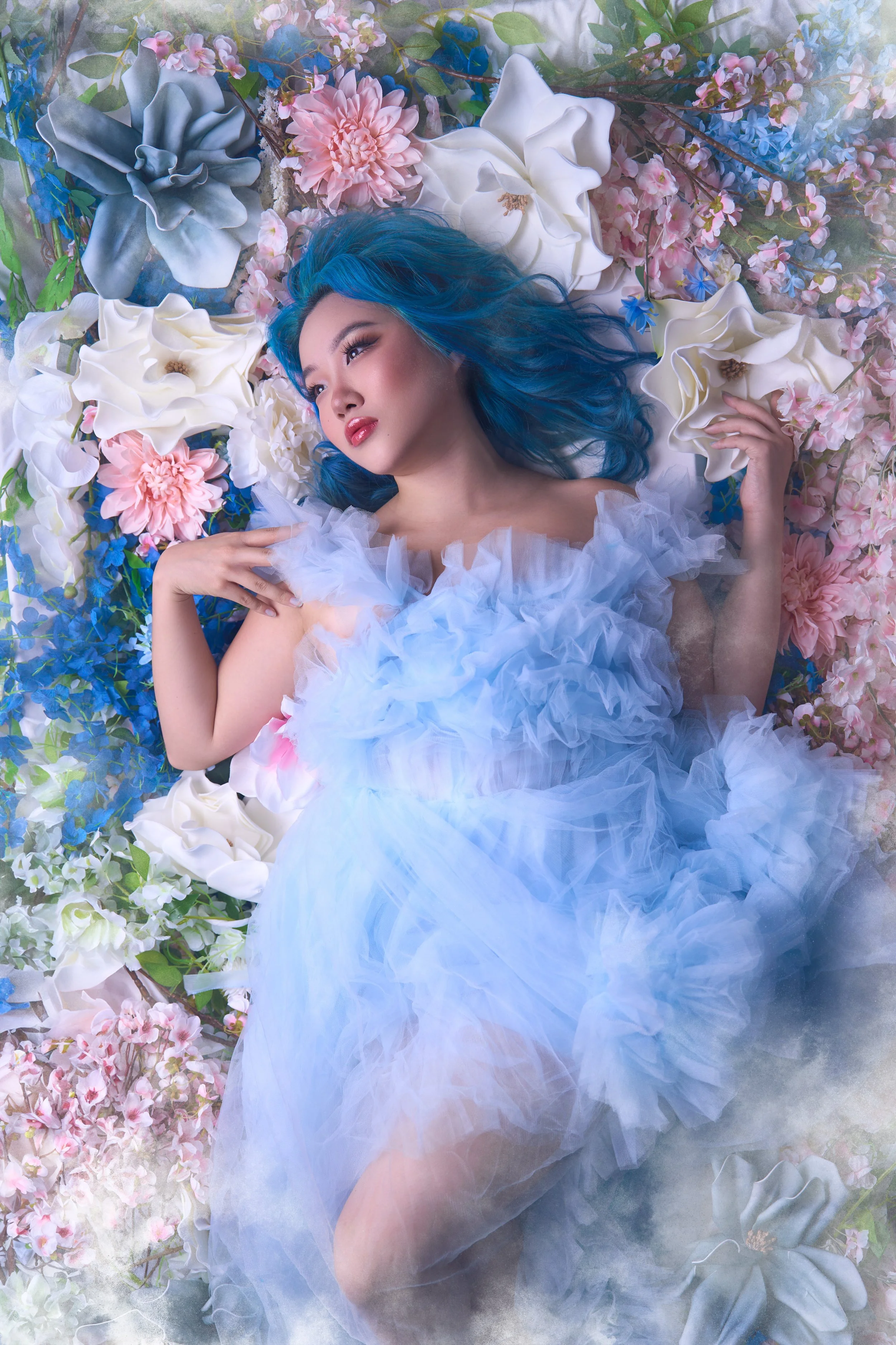 A woman with blue hair lying on a bed of large pink, white, and blue flowers, wearing a light-colored ruffled dress.