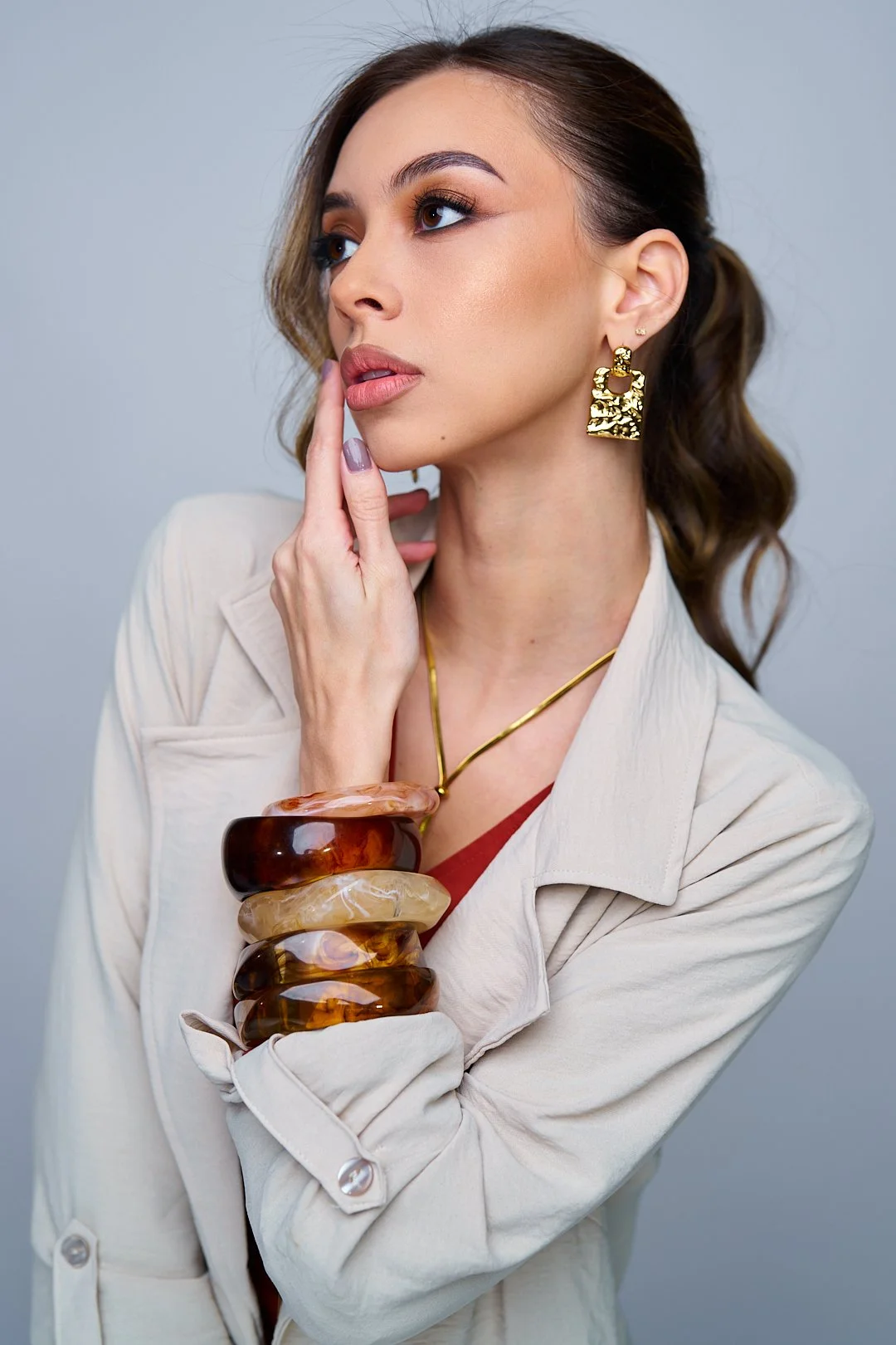 A woman with brown hair styled in loose waves, wearing large gold earrings, a gold necklace, a beige blazer, and multiple chunky amber bangles, touching her lips with her finger.