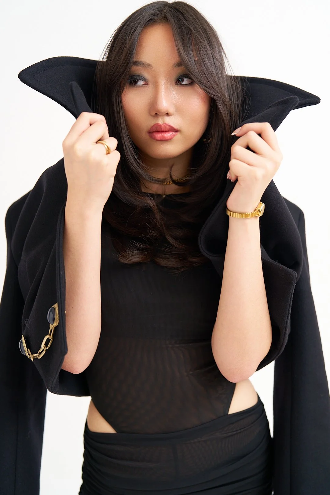 Woman with long dark hair, dressed in black, holding the collar of her coat, with gold jewelry, against a plain white background.