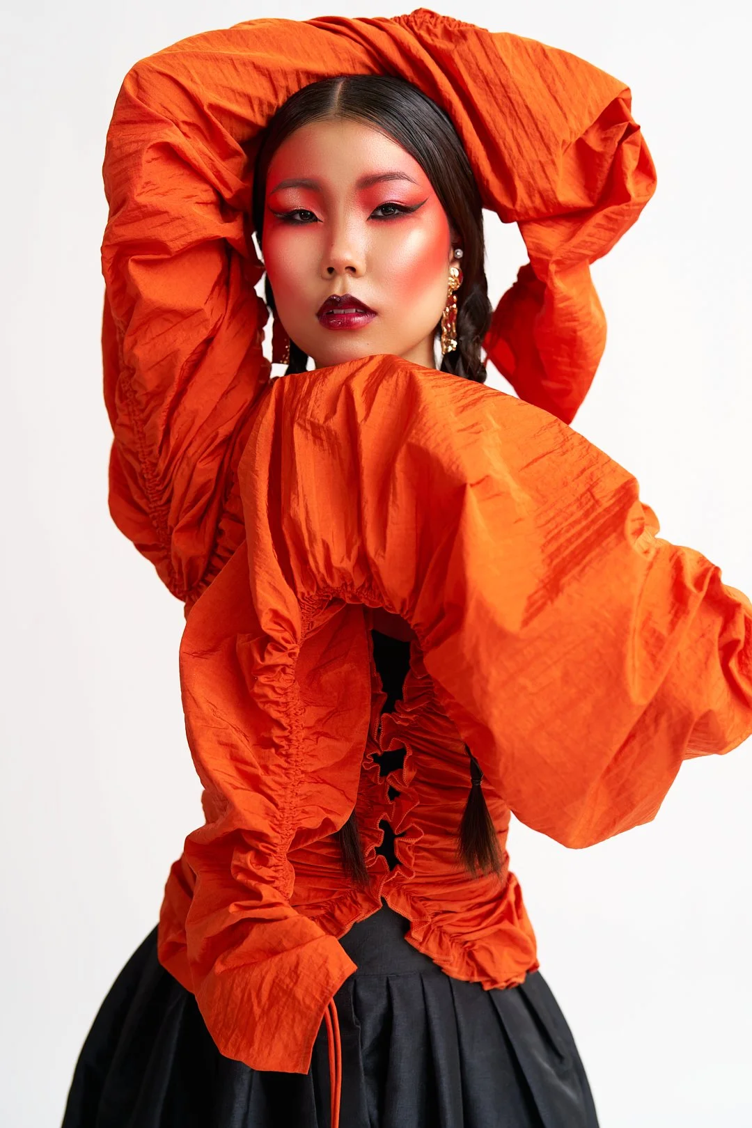 A woman with dark hair styled in braids, wearing bold makeup with red eyeshadow and dark lipstick, poses with one arm raised. She is dressed in an orange, ruched, oversized jacket and a black skirt, with noticeable jewelry including earrings.