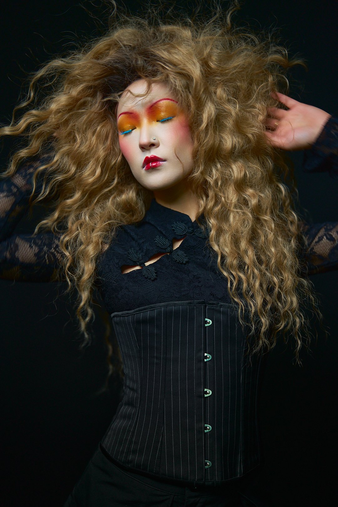 A woman with long, curly blonde hair wearing bold makeup with yellow and orange eyeshadow, blue eyeliner, and red lipstick. She is dressed in a black lace top and a black striped corset, posing with her eyes closed against a dark background.