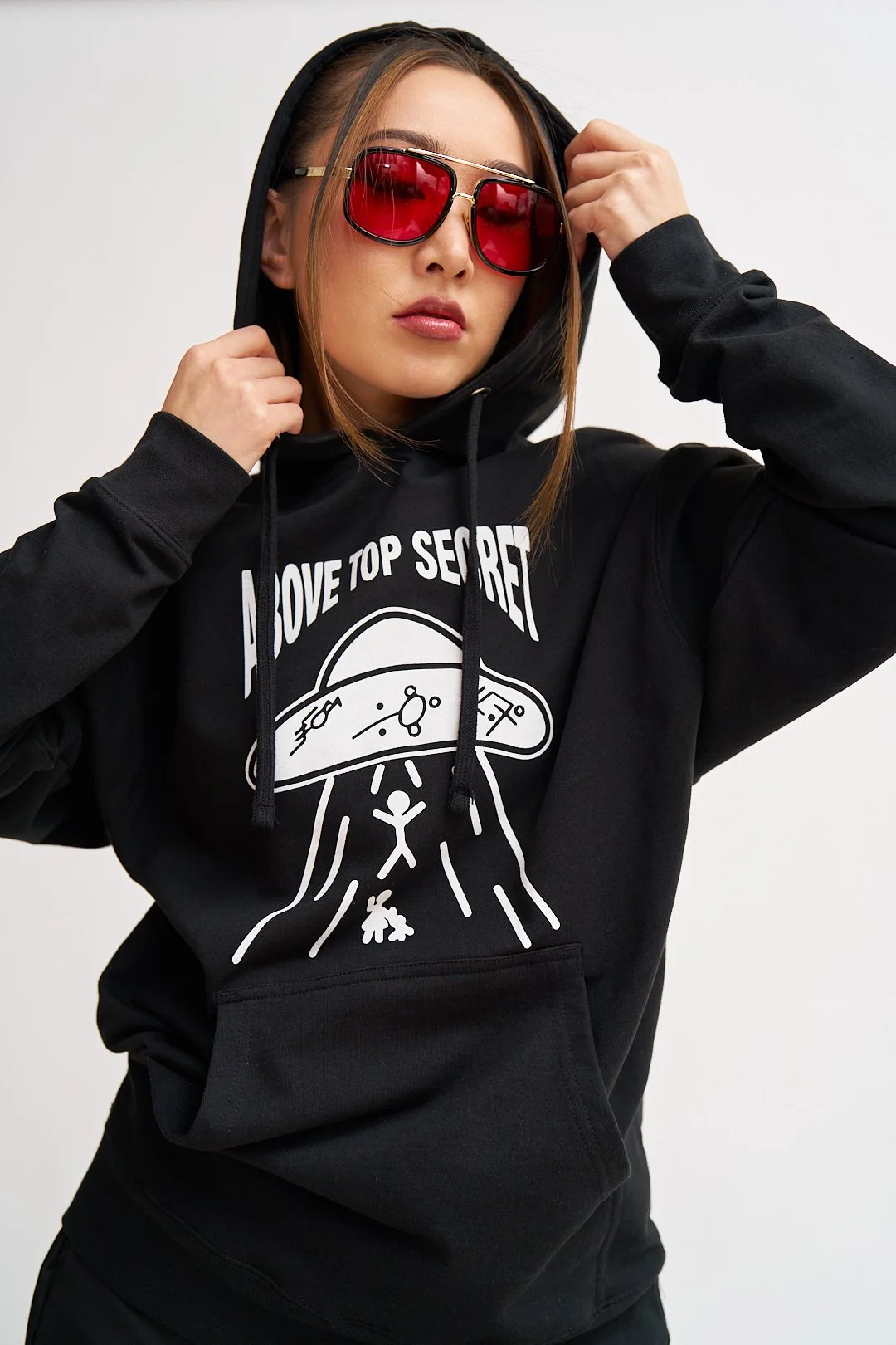 Young woman wearing black hoodie with graphic and text, red sunglasses, and adjusting the hood.