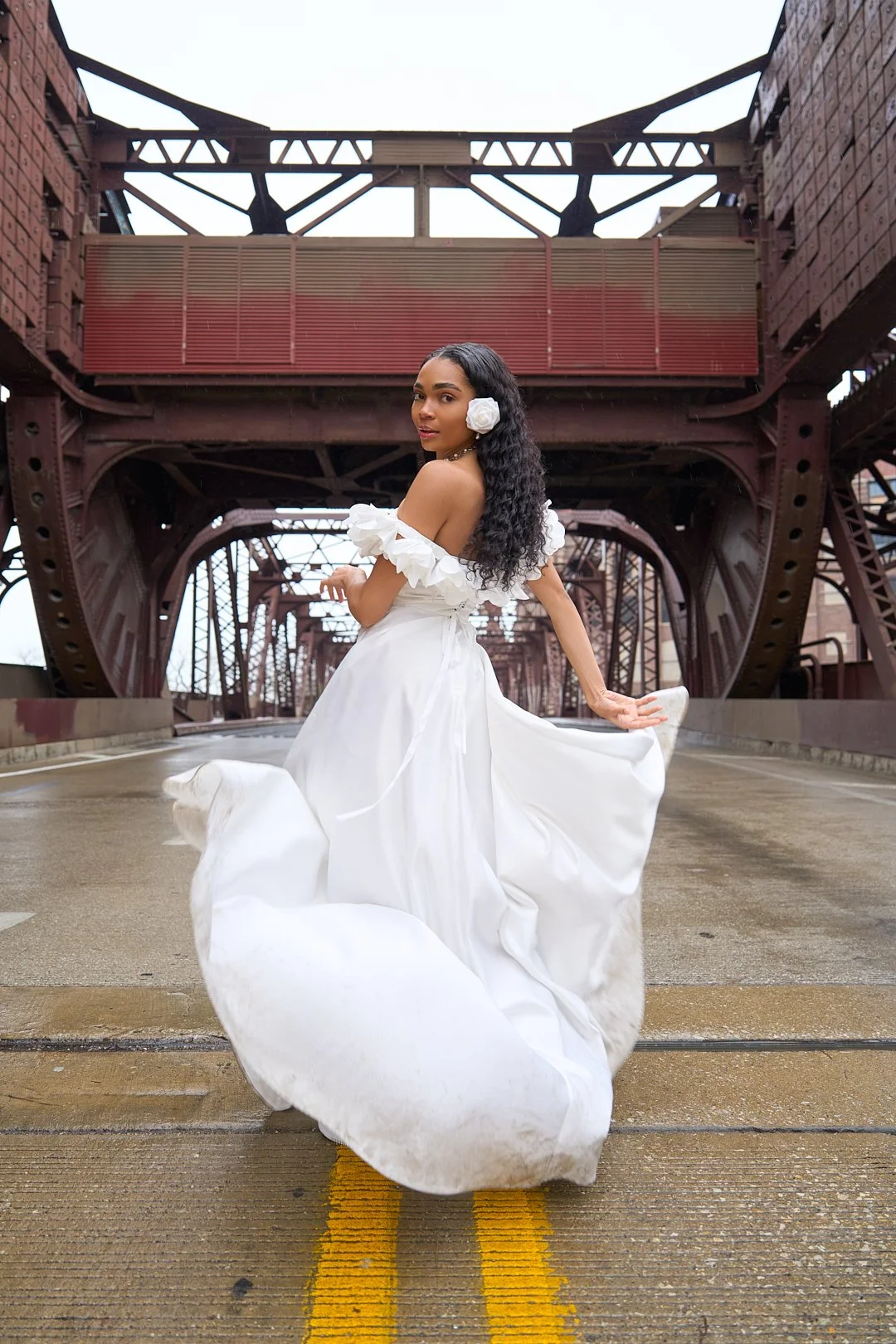 A woman in a white wedding dress with ruffled sleeves and a large rose hair accessory pose on a bridge with industrial architecture.