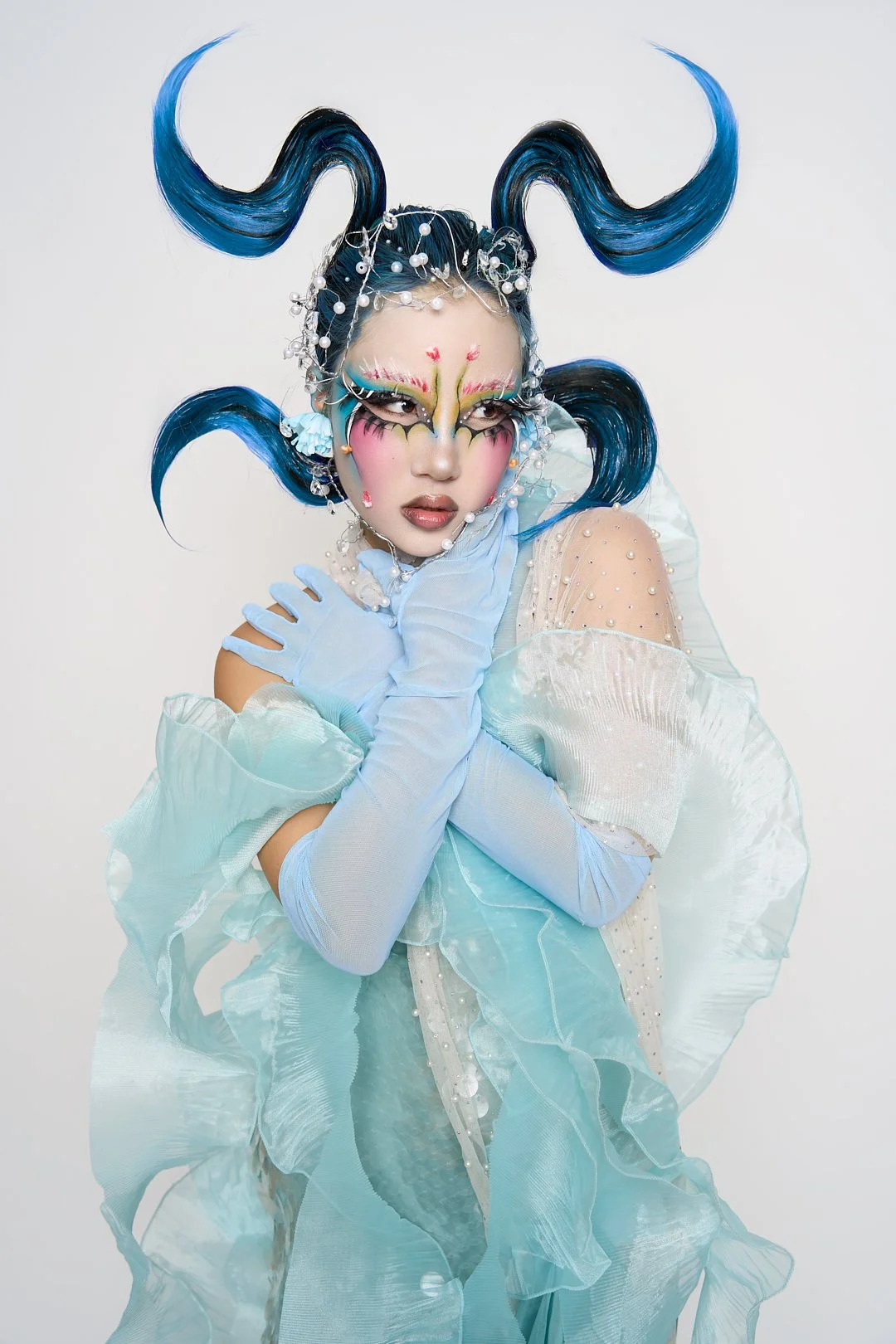 A person with elaborate blue hair styled into large, curled shapes, adorned with pearls and crystals, wearing bold fantasy makeup, a flowing light blue dress with textured layers and pearl embellishments, and light blue gloves, posing against a plain