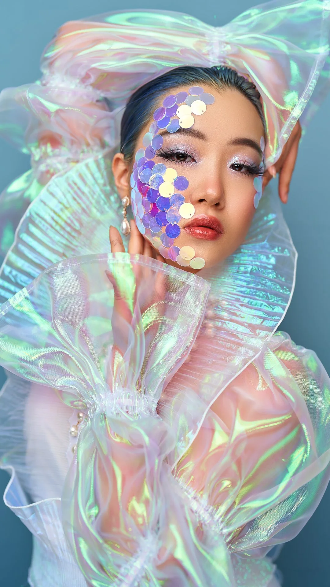 A woman wearing iridescent, holographic clothing with transparent, flowing fabric and sequins on her face, posing with her hand near her face against a light blue background.