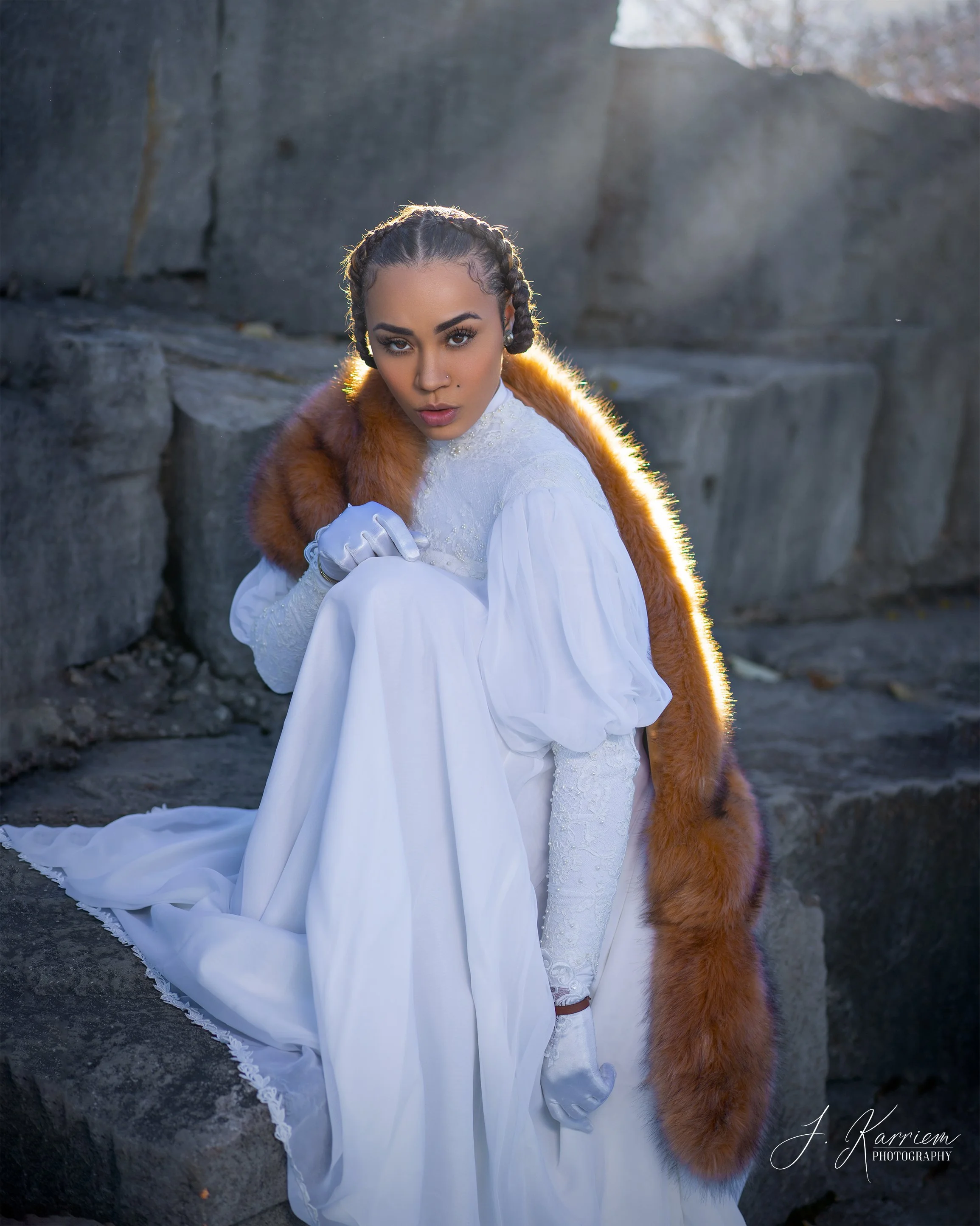A woman in a white vintage dress with lace details and long sleeves, sitting on rocks outdoors in the sunlight, wearing a brown fur stole around her shoulders, with braided hair, gloves, and makeup, looking intently at the camera.