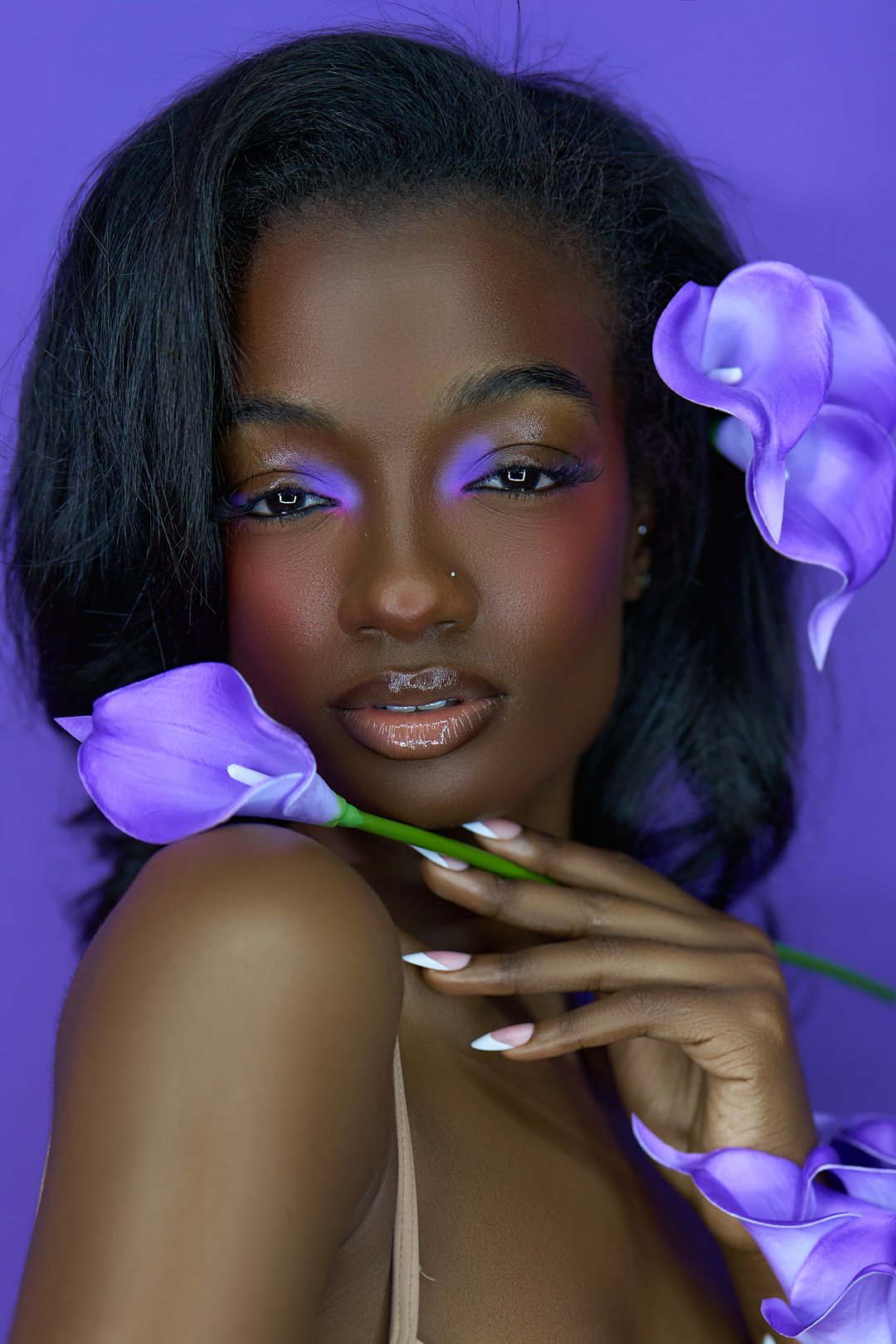 A portrait of a woman with dark skin and black hair, holding light purple calla lilies around her face, against a purple background, with makeup featuring purple eyeshadow and glossy lips.