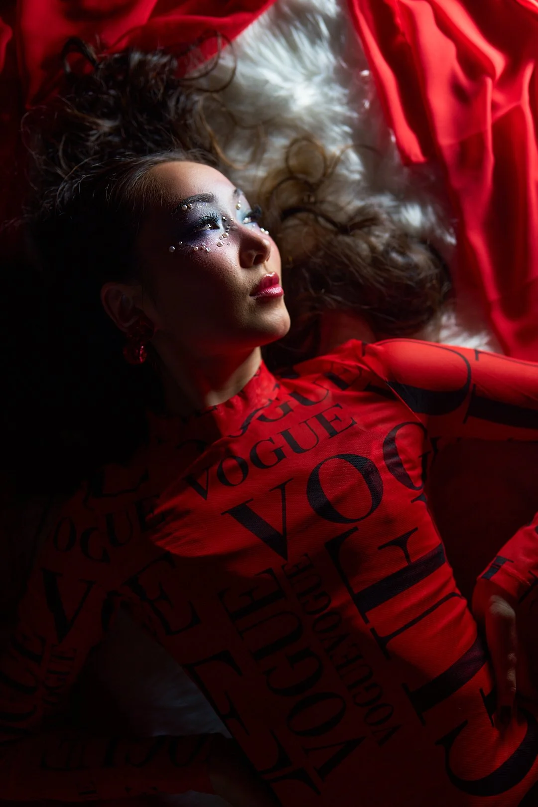 A woman lies on her back on a fluffy surface, wearing a red dress with black text and looking upward. She has elaborate makeup with sparkles around her eyes and dark lipstick, and long wavy hair spread around her.