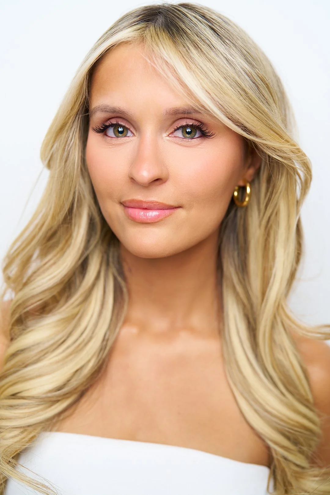 Close-up portrait of a blonde woman with wavy hair, wearing hoop earrings and a white top, against a white background.