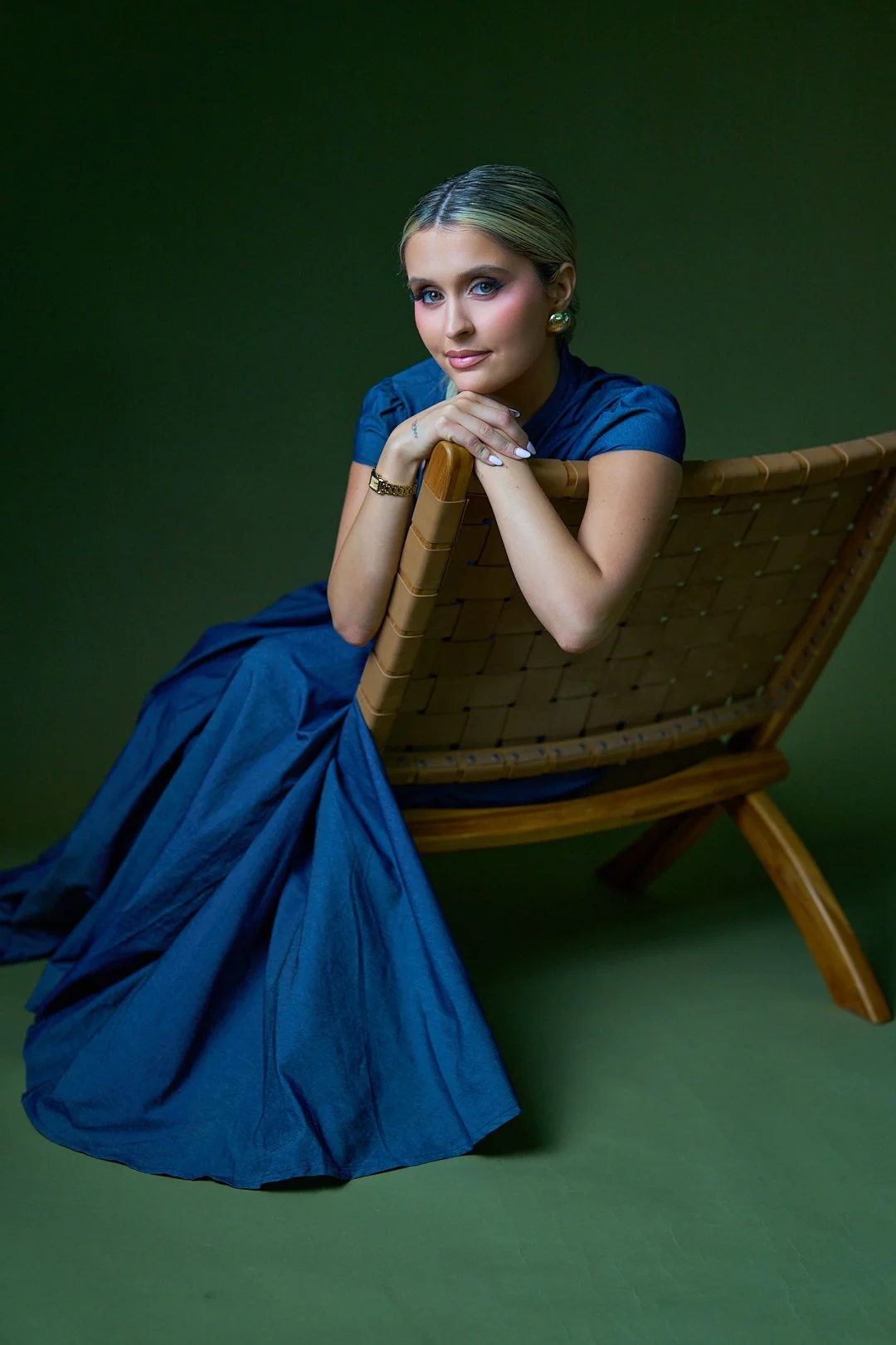 A woman in a blue dress sitting on a wooden chair with a green background, resting her chin on her hands.