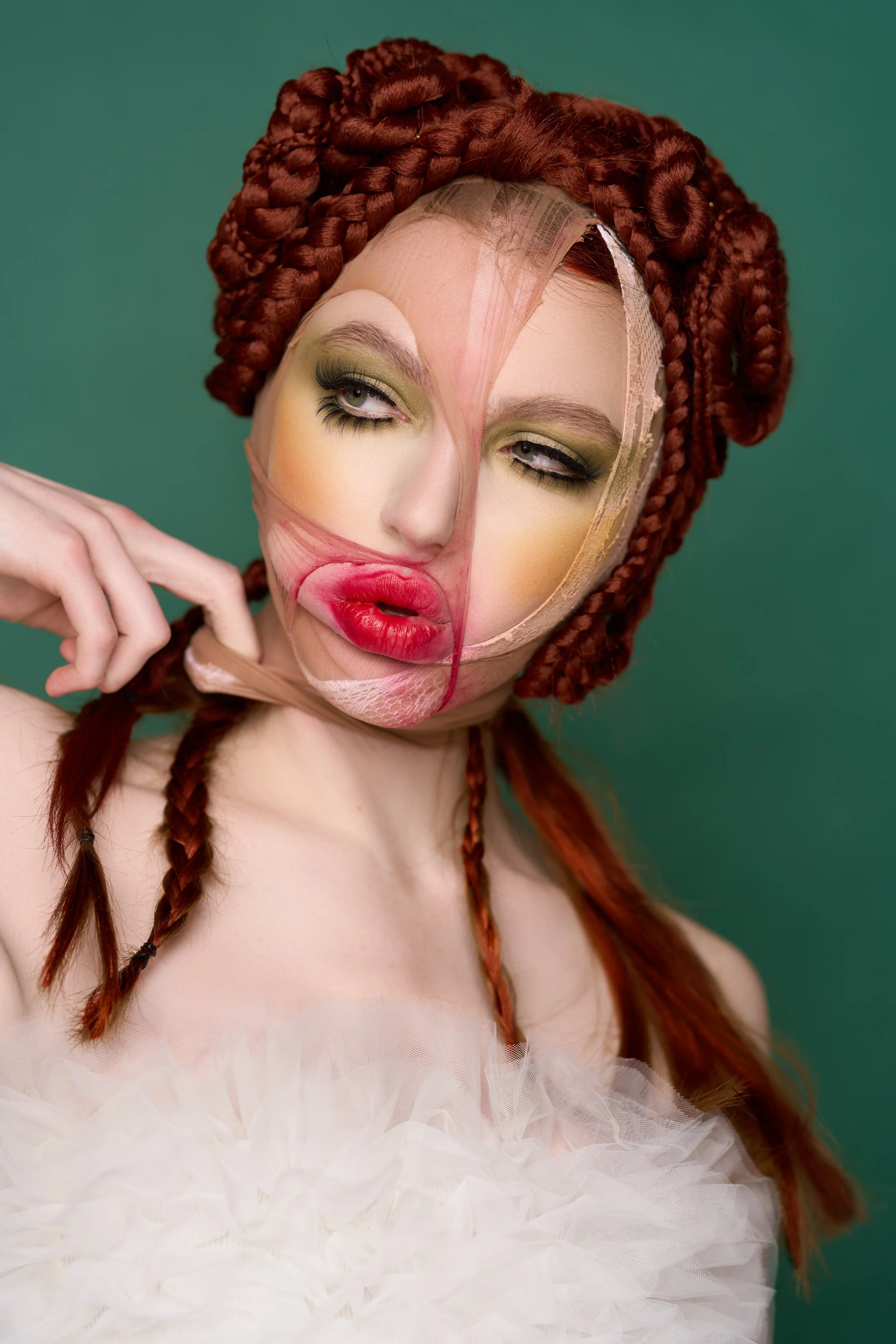 A woman with red braided hair is wearing yellow eyeshadow and lipstick. Her face appears partly covered with distorted, blurry promotional face mask, creating a surreal effect.