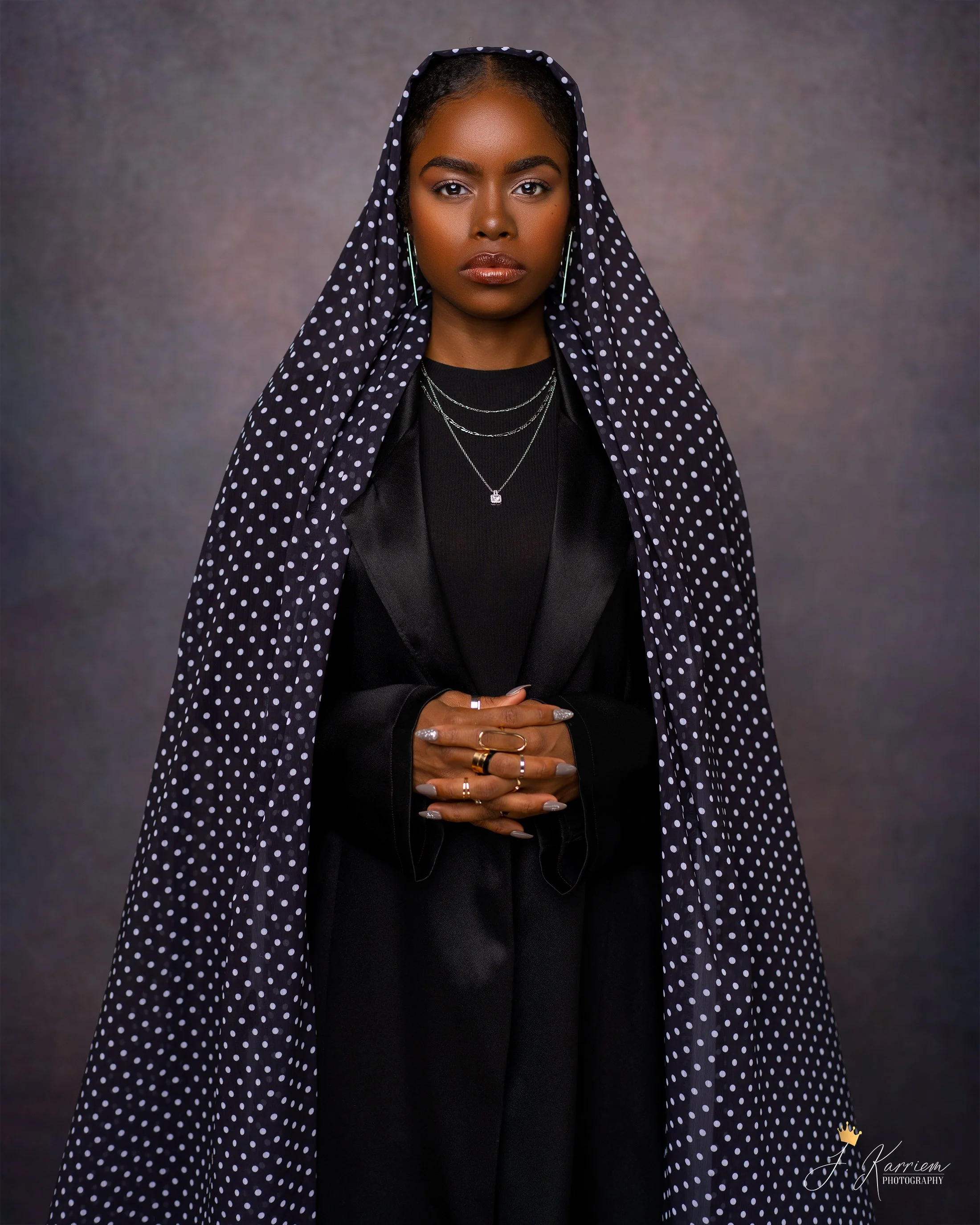 A woman with dark skin and short hair wearing a black outfit with layered necklaces, rings, and earrings is standing with a serious expression against a textured gray background. She has a black polka dot fabric draped over her head and shoulders.