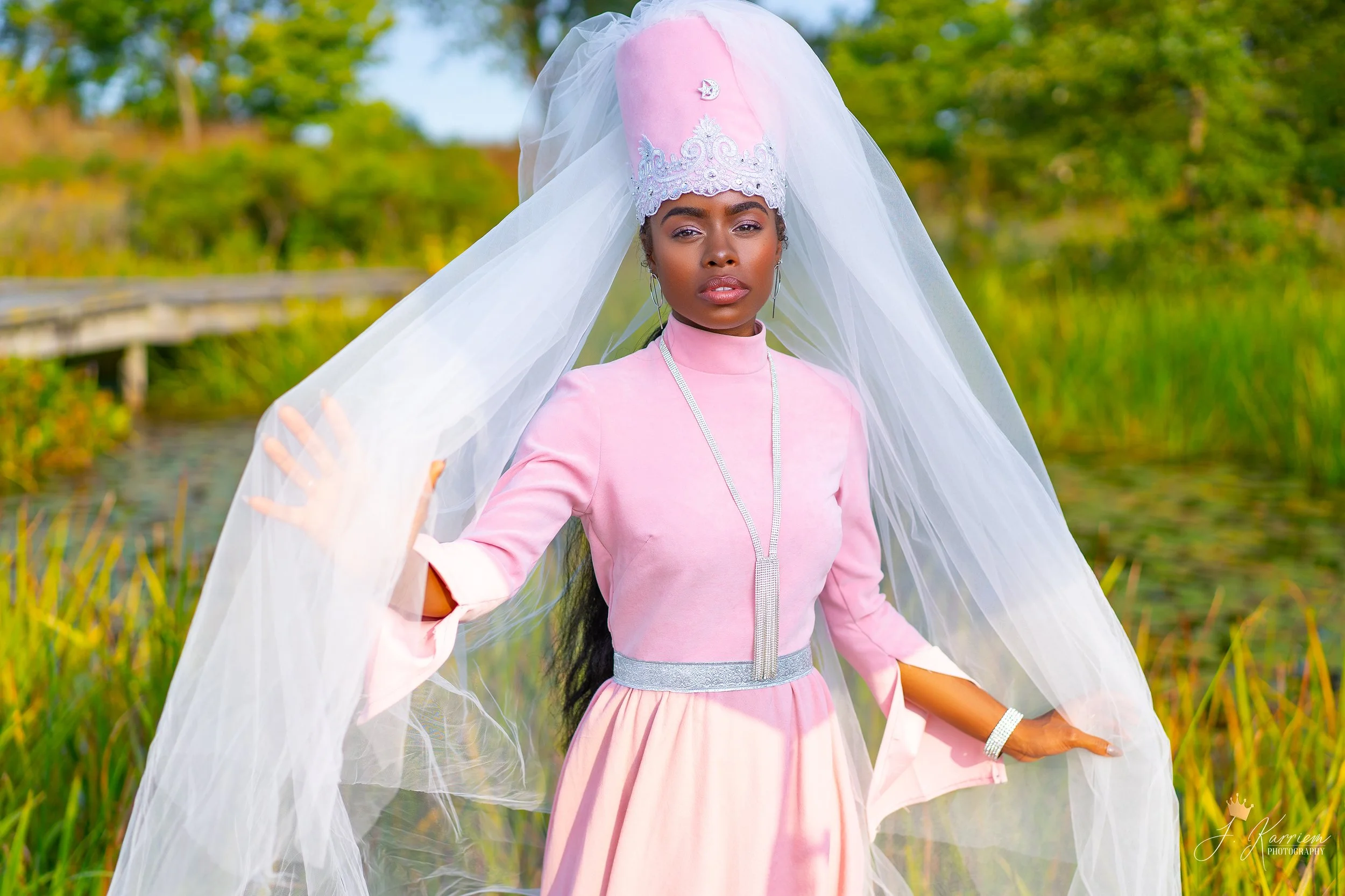 A woman dressed in a pink dress with a long veil and a tall pink hat with lace and jewel embellishments, standing outdoors by a body of water with greenery in the background.