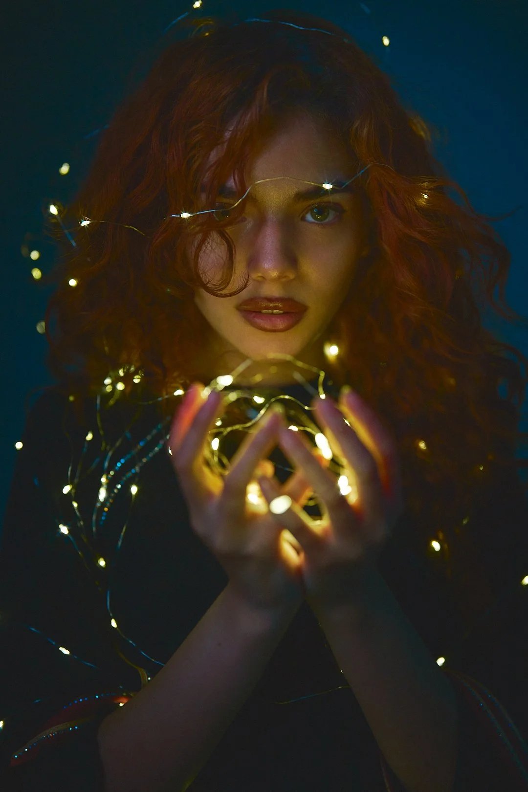 A woman with curly red hair holding a gold string of fairy lights close to her face, with some lights wrapped around her hair and hands, against a dark background.