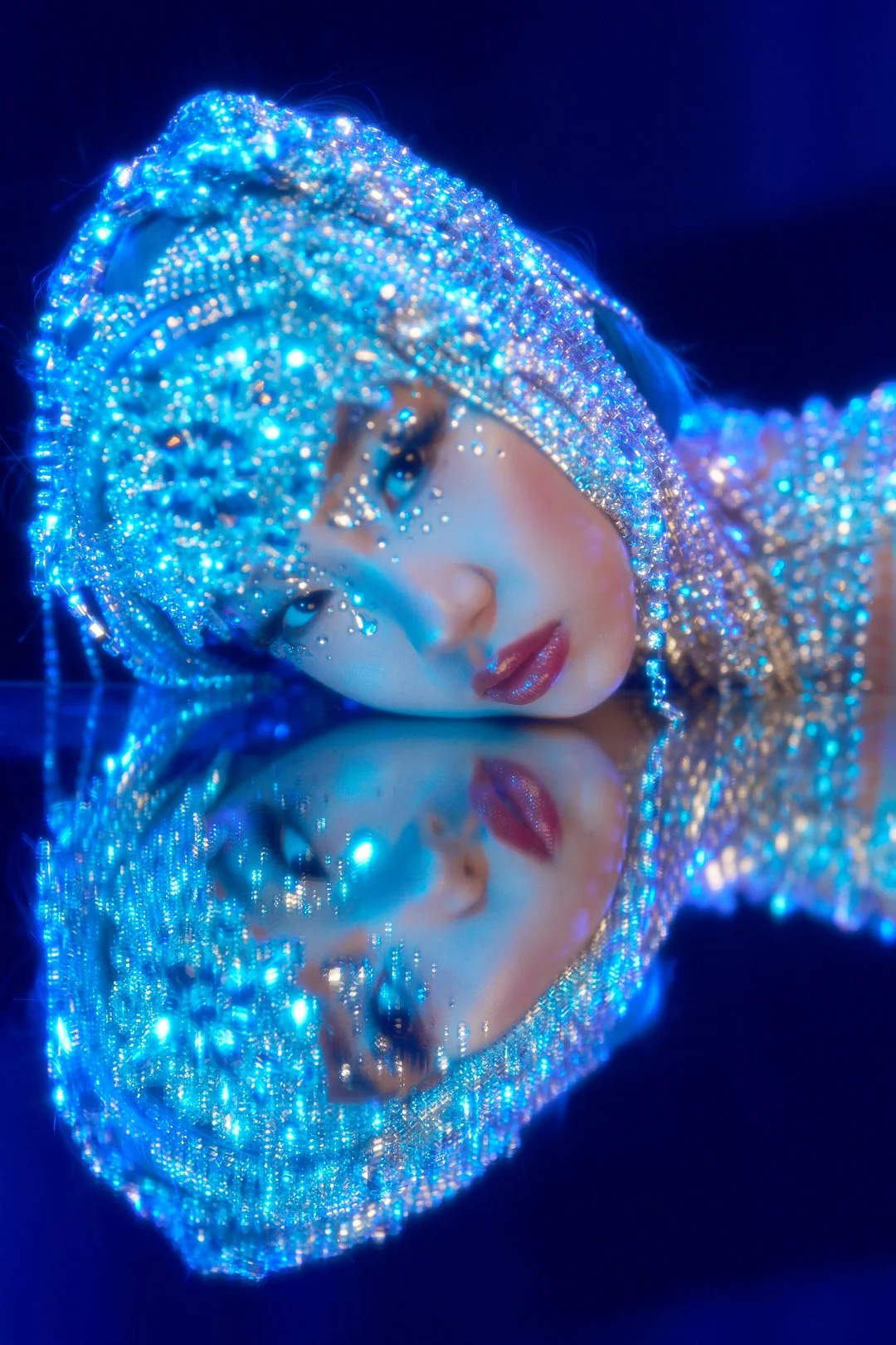 A woman wearing a headpiece and dress adorned with blue and silver lights, with her reflection on a glossy surface.