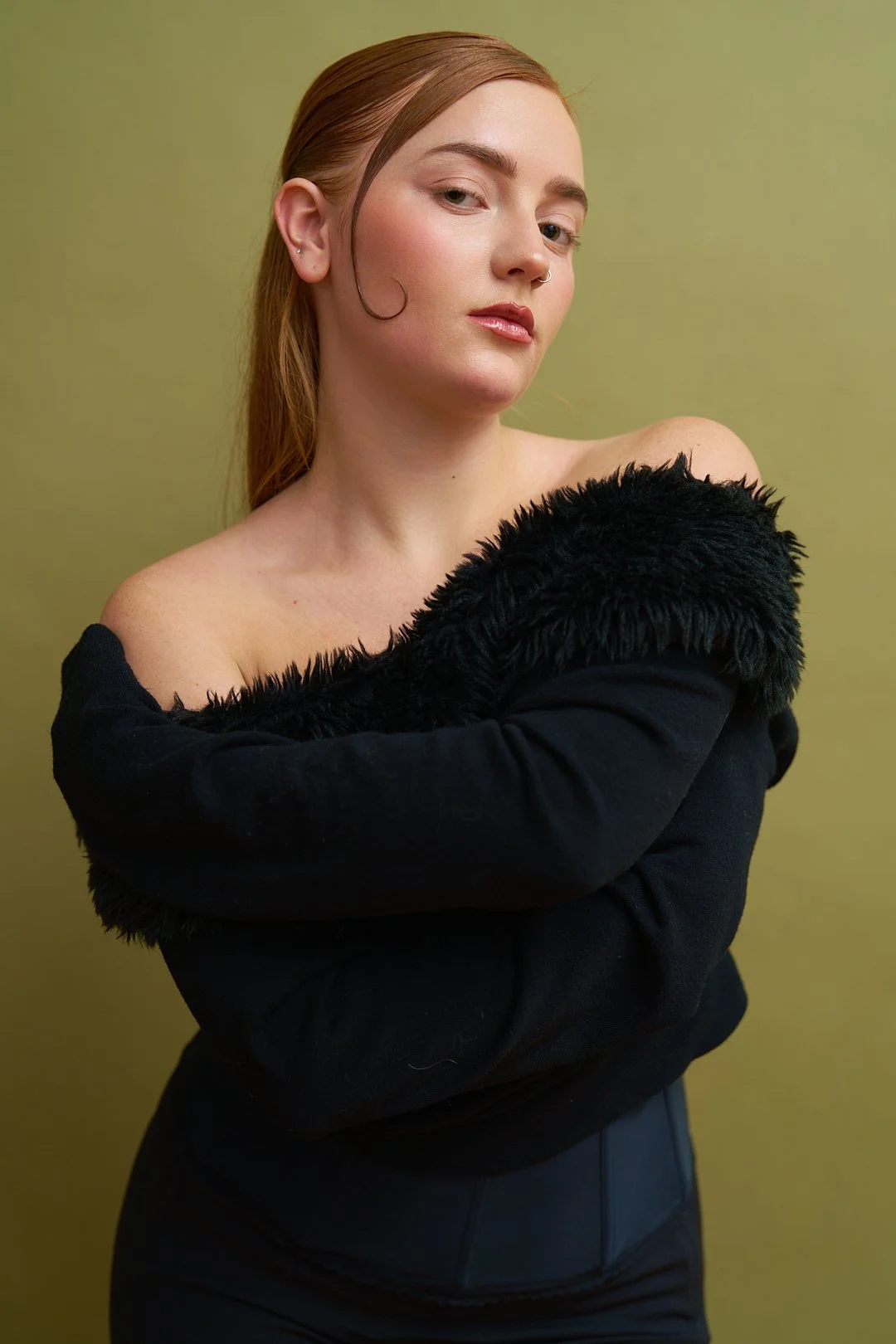 A woman with red hair, wearing a black off-shoulder top with a fuzzy collar, stands against a plain light green background.