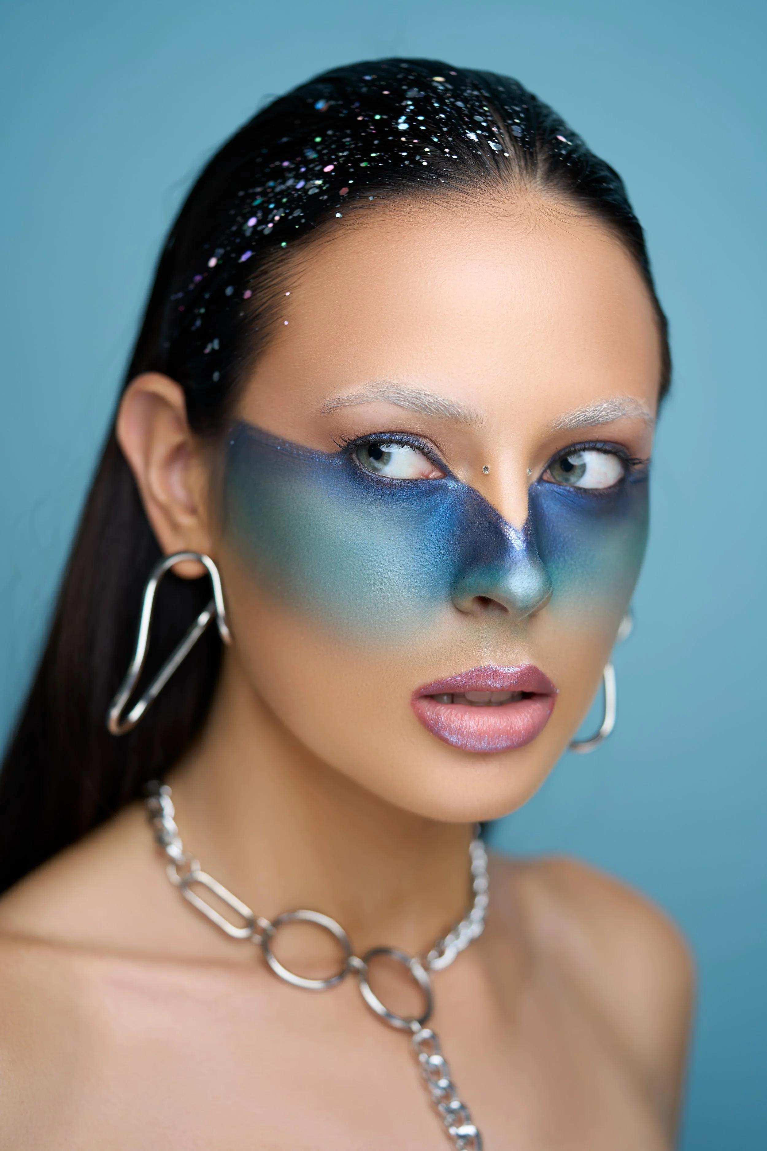 Woman with metallic blue, green, and purple face paint, silver jewelry, and glitter in her hair against a blue background.