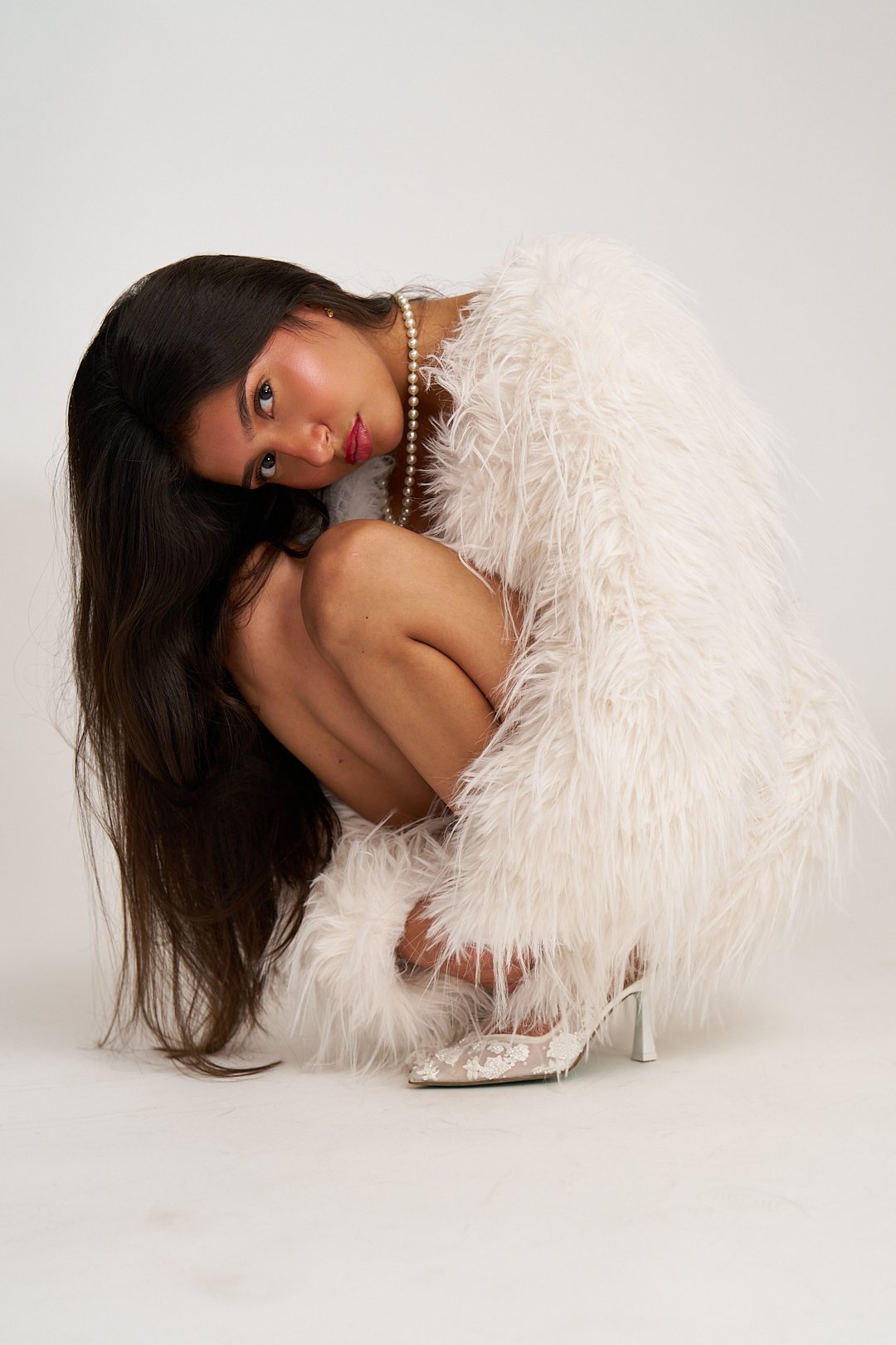 A woman with dark hair and makeup, wearing a pearl necklace, a white feathered boa, and patterned high heels, is crouching in a curled pose against a plain white background.