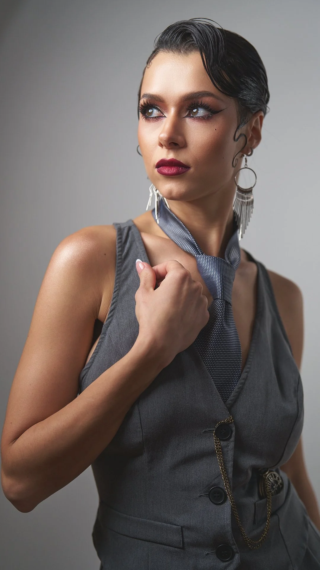 A woman with dark hair styled in a vintage wave, wearing a gray sleeveless dress with black buttons, a blue and gray neck scarf, large hoop earrings with fringe, and makeup with bold lipstick, looking to the side.