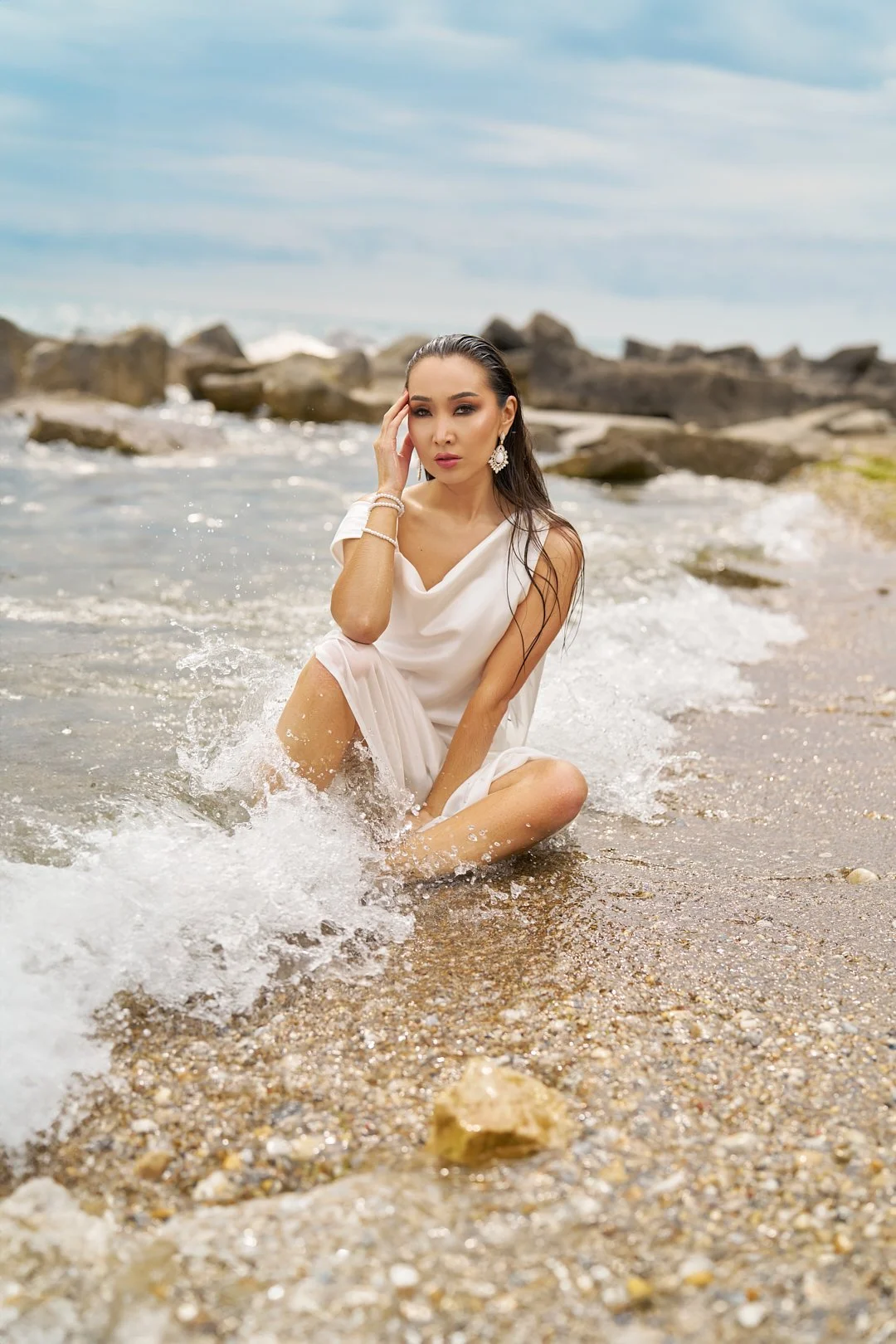 A woman sitting in shallow ocean water near rocks on the beach, wearing a white dress and jewelry, posing with one hand on her face and looking at the camera.