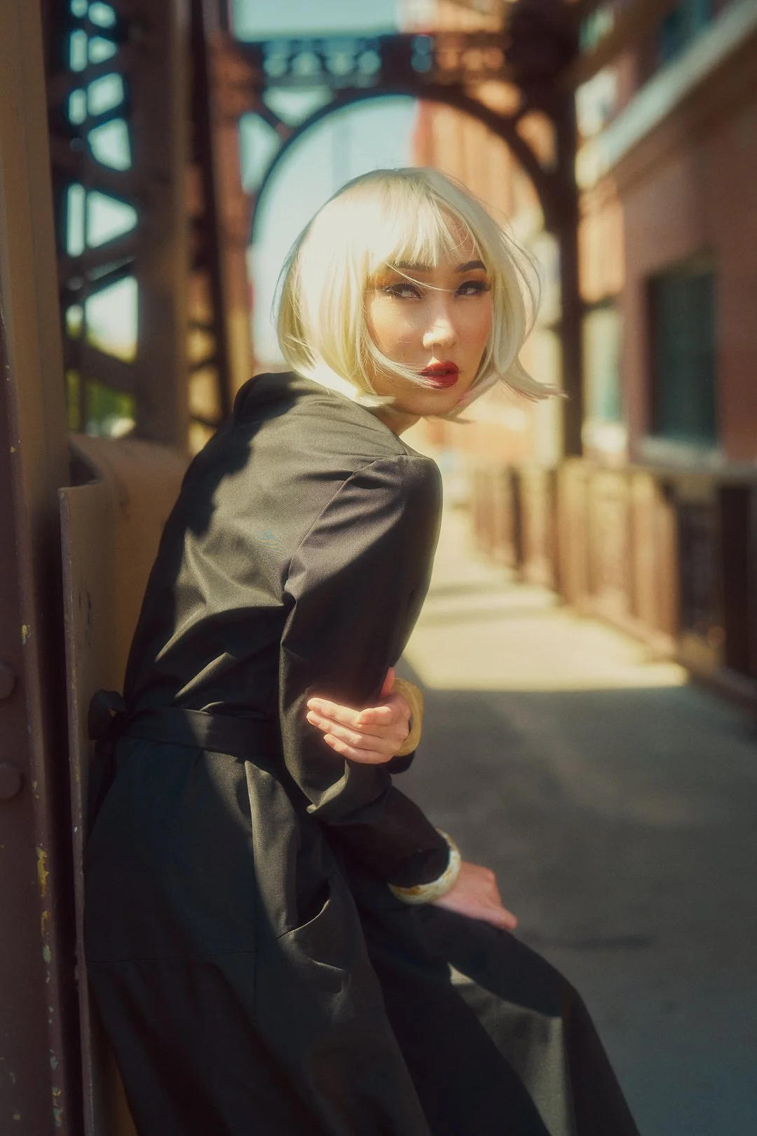 A woman with platinum blonde hair and red lipstick sitting outdoors under an iron bridge on a sunny day, wearing a black satin blazer.