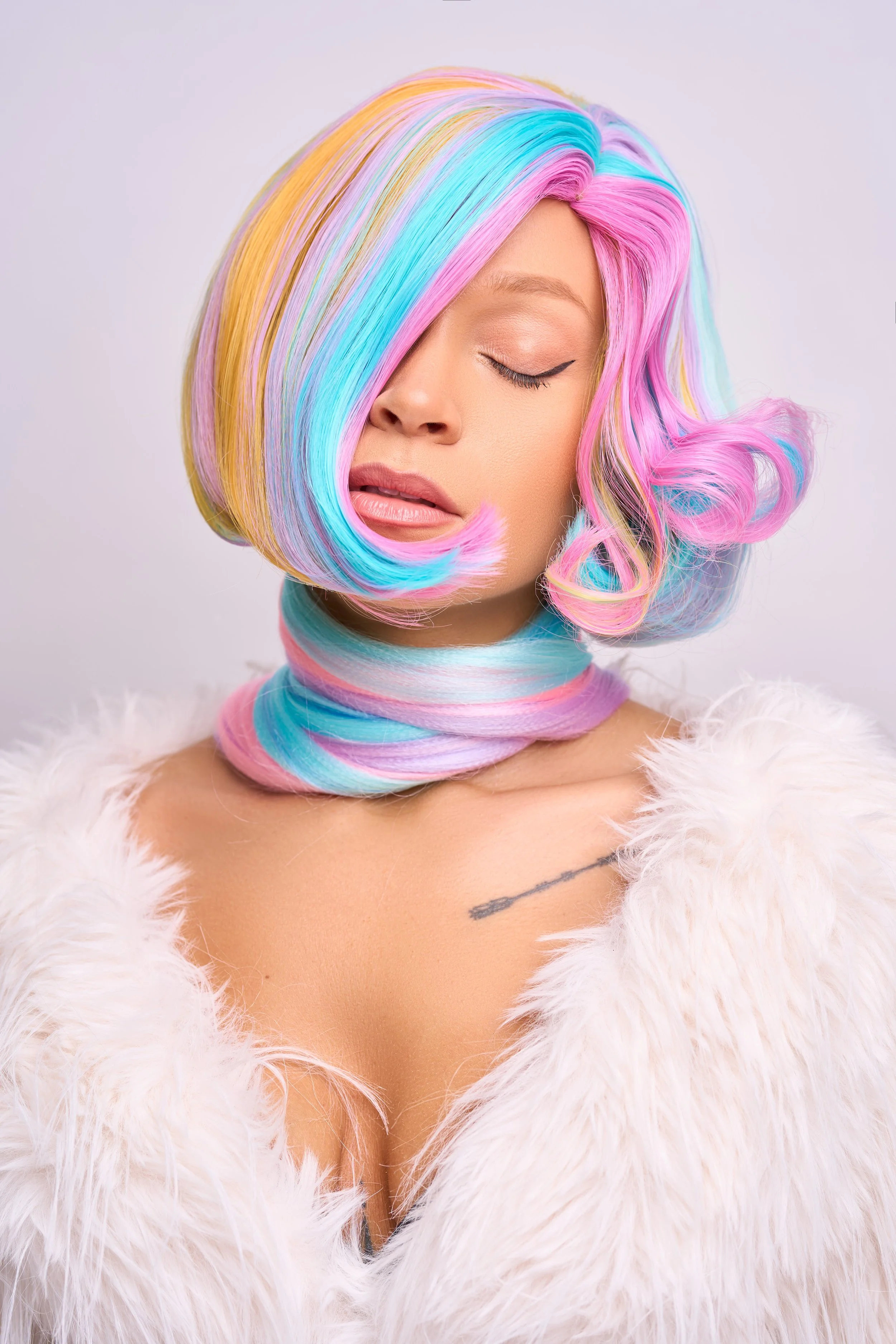 A woman with multicolored, pastel rainbow hair styled in large curls, wearing a fluffy white coat, with a pastel-colored scarf wrapped around her neck and a small tattoo visible on her chest, against a plain light background.