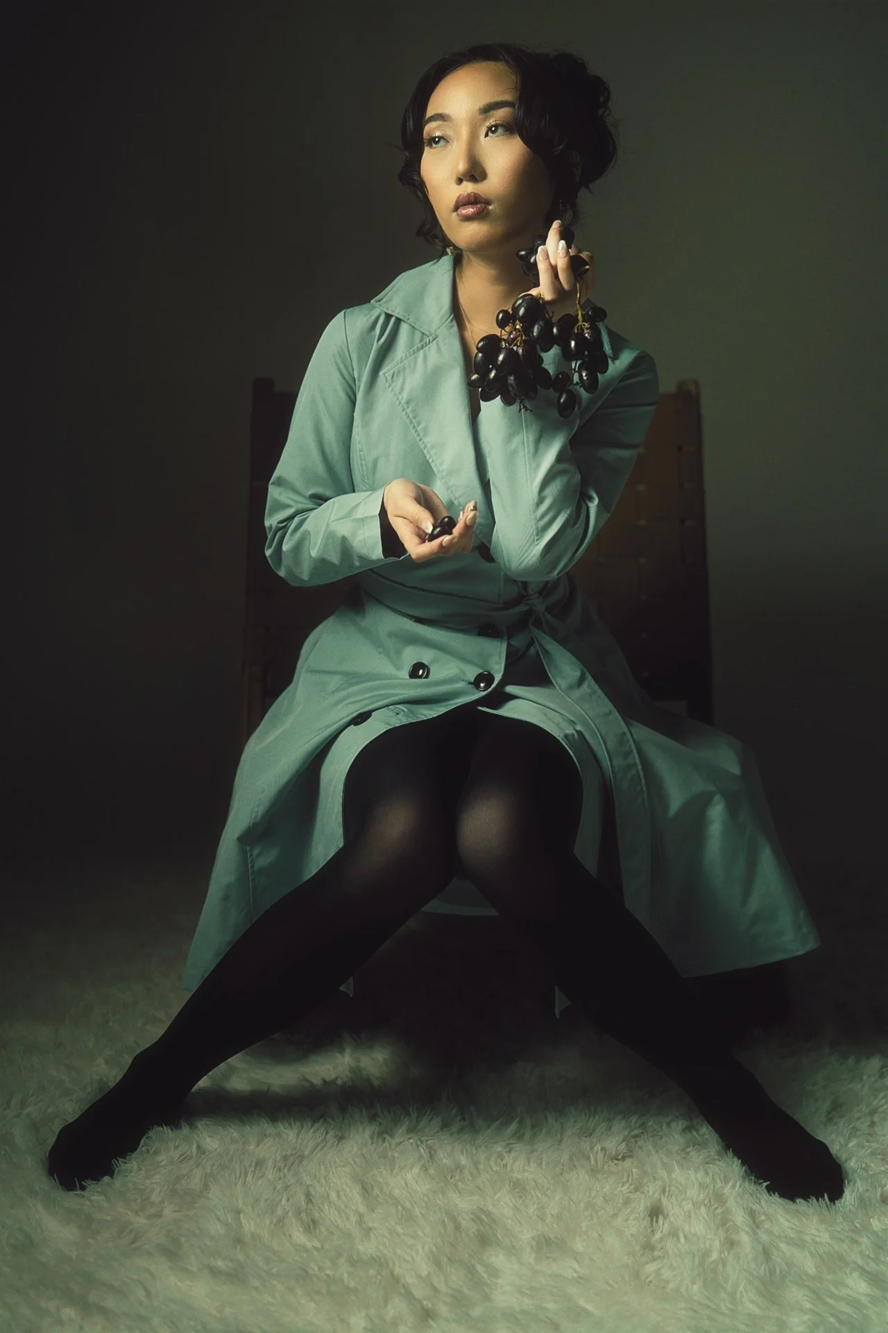A woman in a light green trench coat is sitting on the floor with her legs apart, wearing black tights. She holds a bunch of black grapes near her face with her right hand and a single grape in her left hand. She has short, curly dark hair and a seri