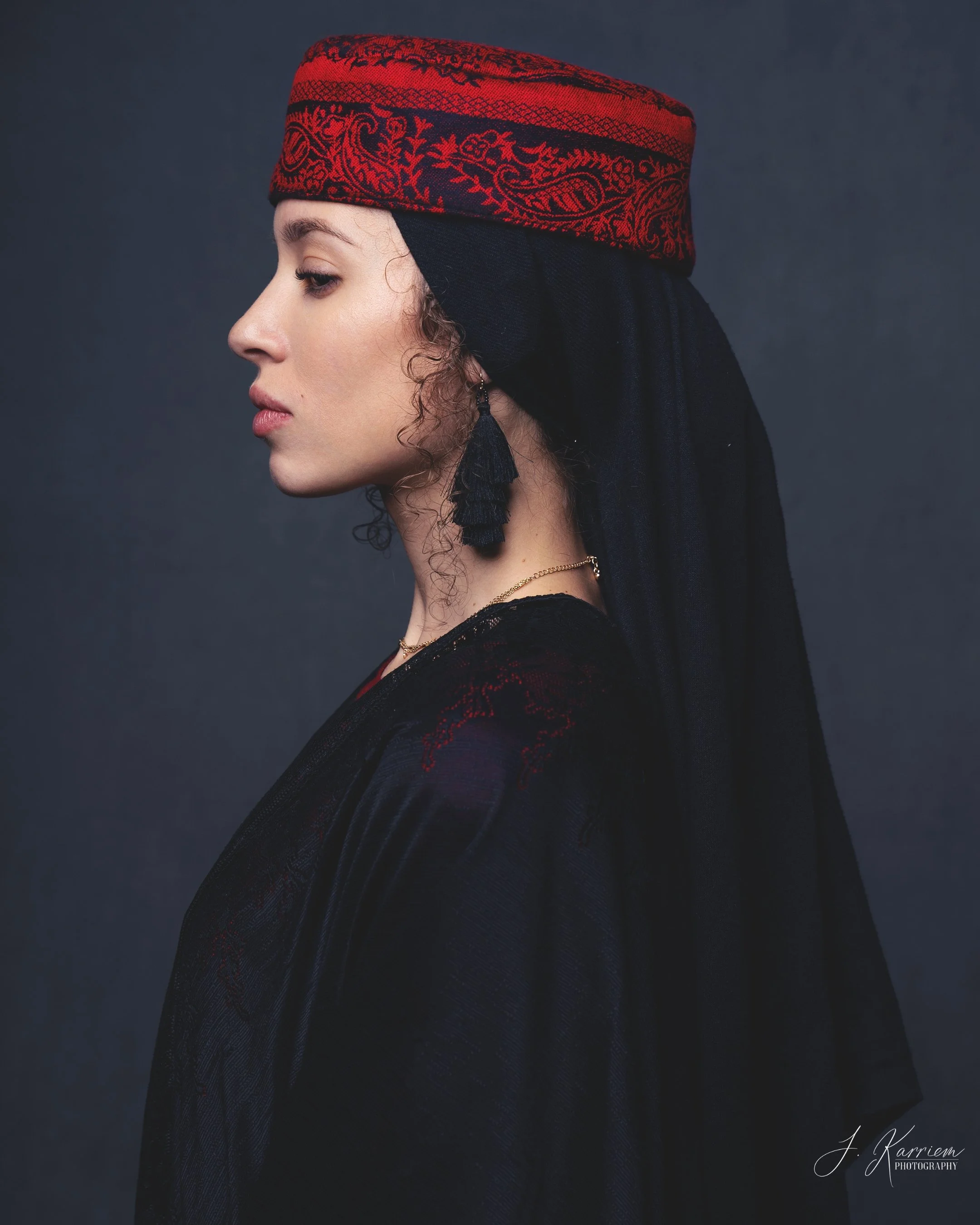 Profile of a woman wearing a red and black embroidered headwrap, black scarf, and black earrings with tassels, against a dark background.