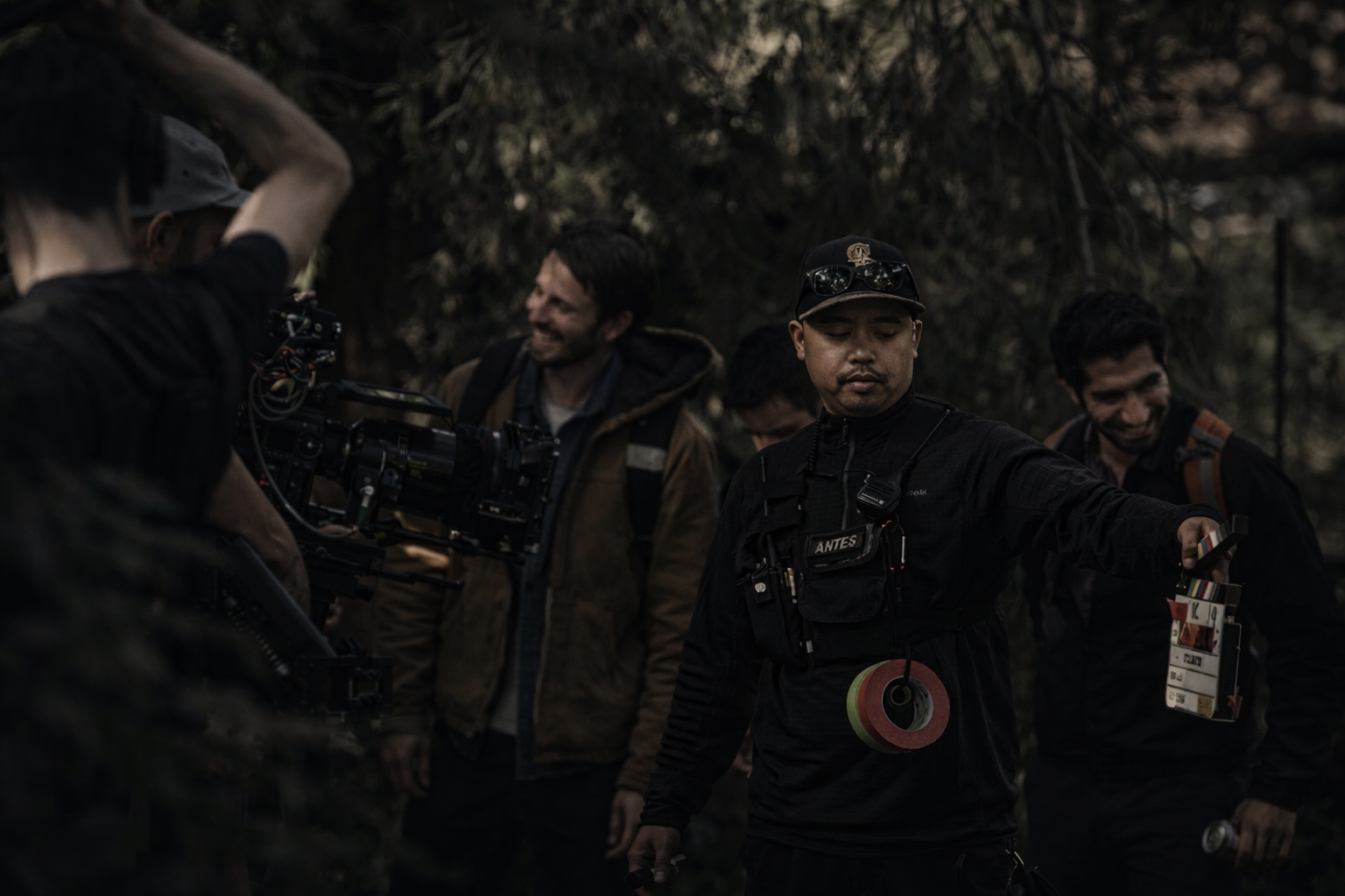 A group of people working on a film set outdoors, with one person holding a clapperboard and another adjusting a device, surrounded by trees in a dimly lit environment.