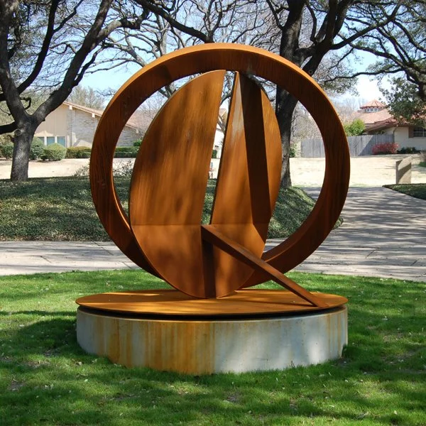 A wooden art sculpture resembling a clock with a circular frame and intersecting triangular shapes, situated on a concrete base in a park-like setting with trees and houses in the background.