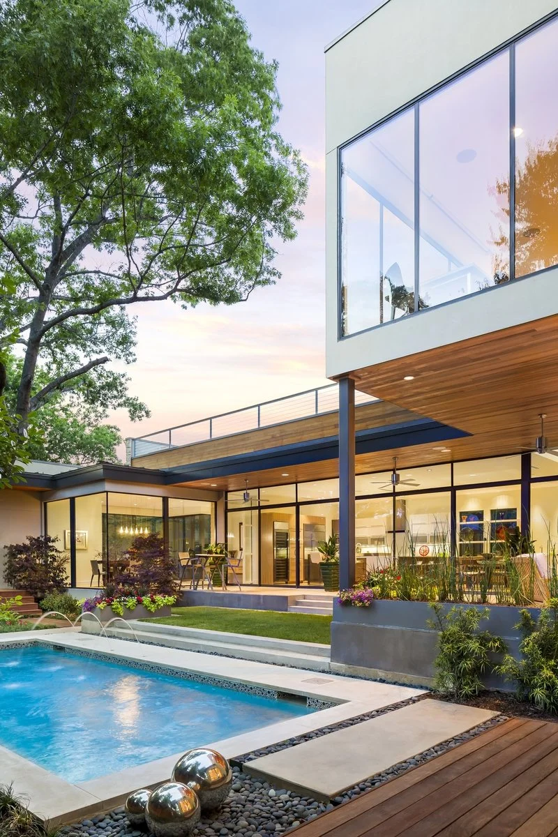 Modern house with large glass windows, outdoor pool, and a patio area with furniture, surrounded by greenery and trees at sunset.