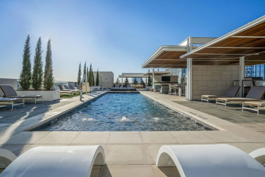 Rooftop pool area with lounge chairs, tall trees, and shaded seating area under steel and wooden pergolas, on a sunny day.