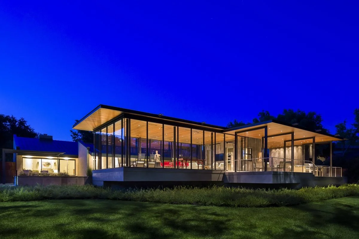 Modern house with glass exterior walls illuminated at night, featuring multiple levels and open-concept living spaces, surrounded by a grassy lawn and trees in the background.