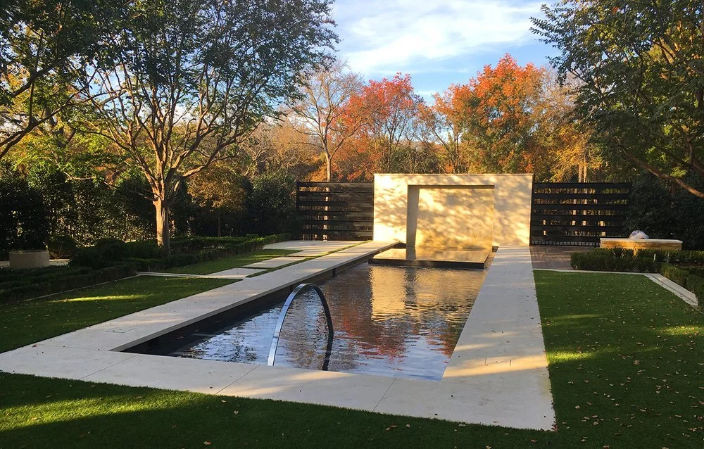 Contemporary backyard with a narrow reflecting pool, white stone walkway, and minimalist sculpture backdrop surrounded by trees with autumn foliage.