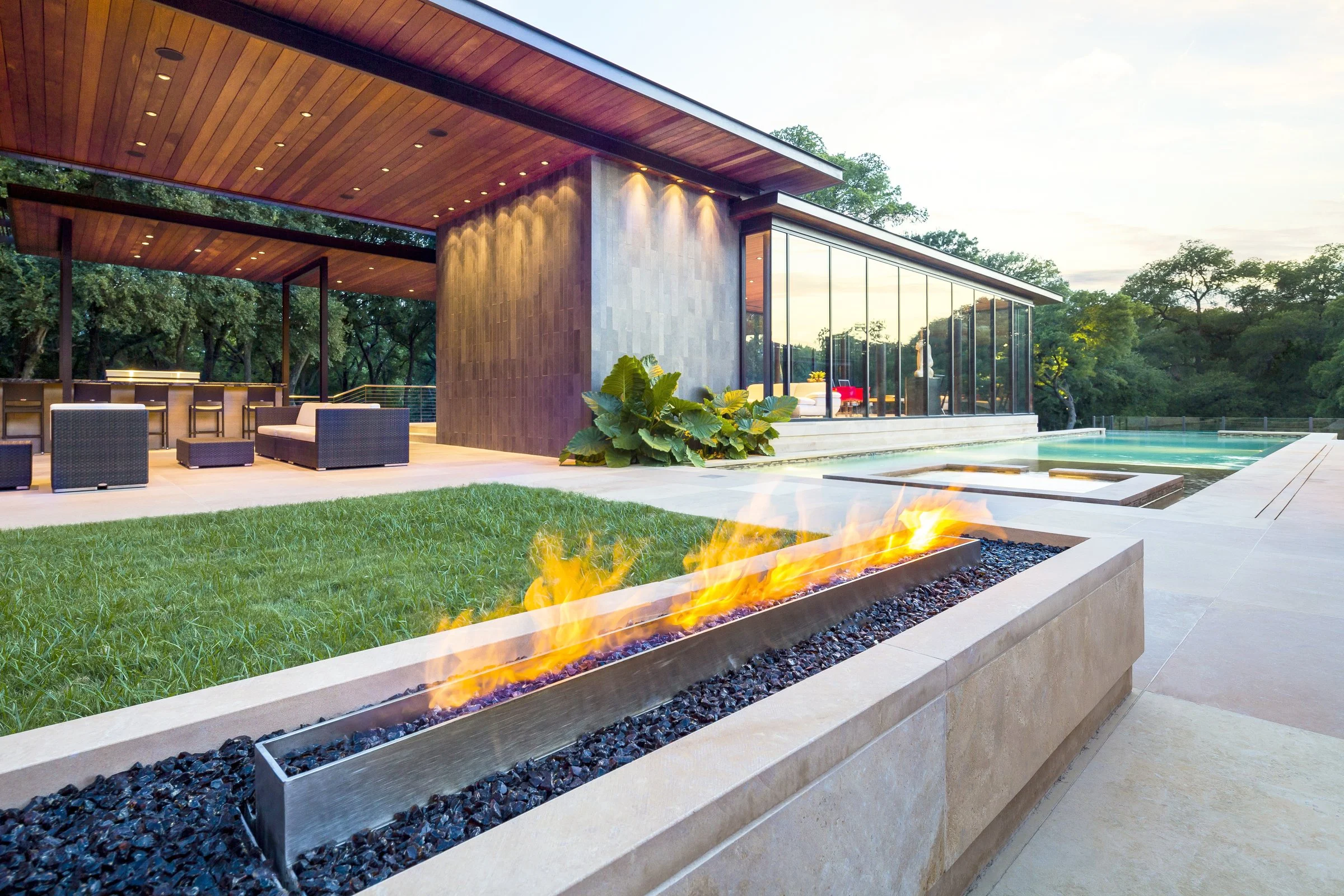 Modern house with a pool and a fire feature in a landscaped yard with trees in the background.