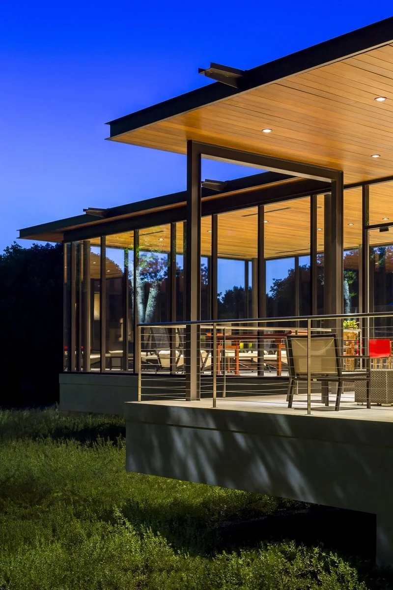 A modern pool house with large glass windows and a wooden roof, illuminated at dusk, with a balcony overlooking a grassy yard.