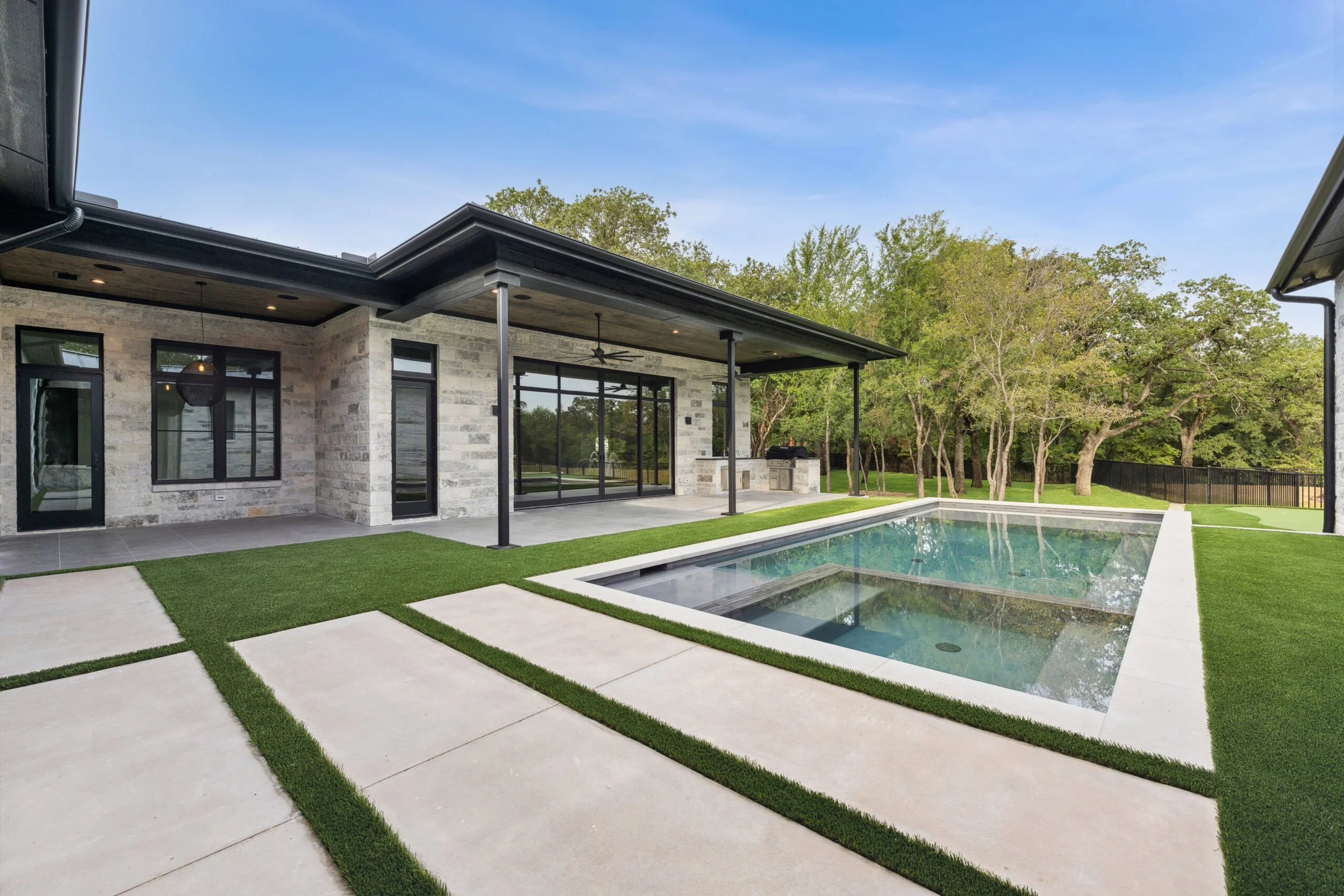 Backyard with a modern house featuring gray brick exterior, large windows, and a covered patio area. In the yard, there is a rectangular swimming pool with a built-in spa, surrounded by well-maintained grass and concrete pathways. Trees are in the ba