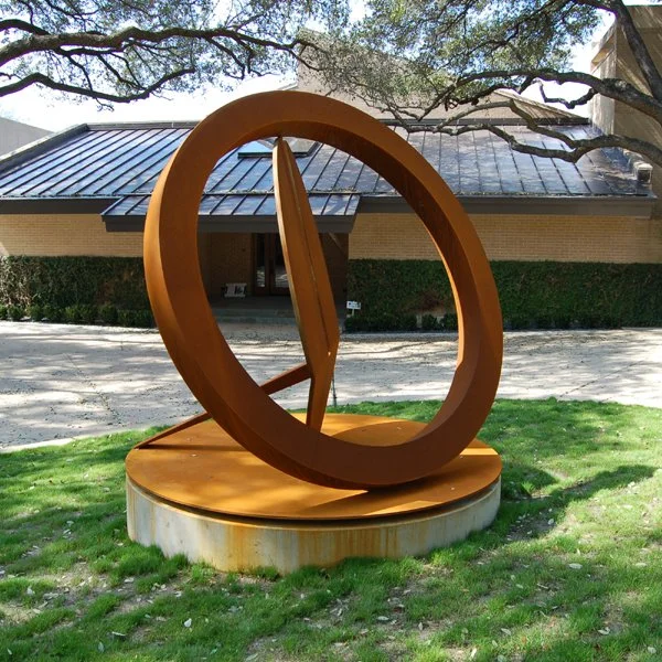 A large modern metal sculpture with circular and triangular shapes, painted in a rust color, situated outdoors on a grassy area with trees and a building in the background.