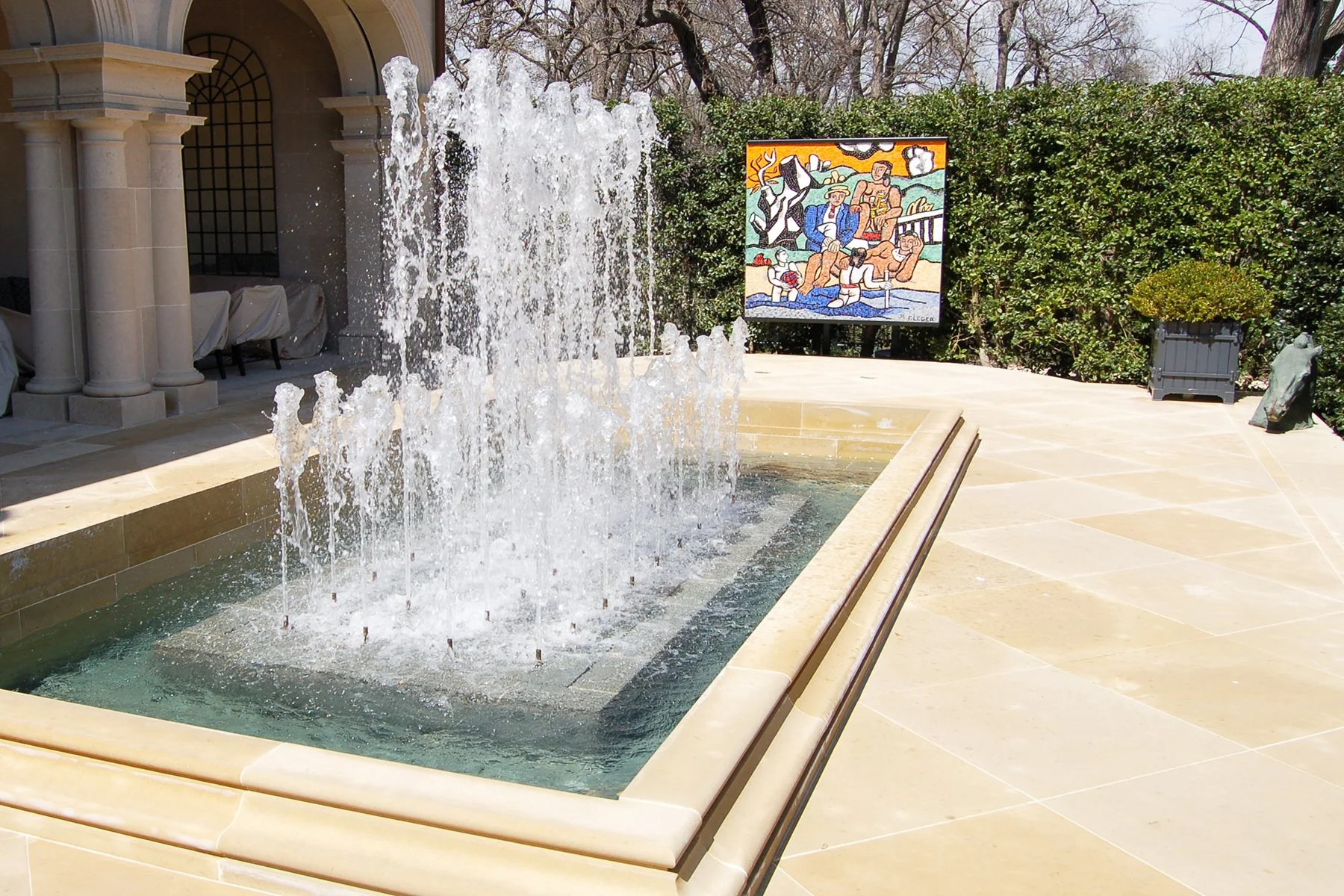 A fountain with water jets in an outdoor courtyard, with a colorful mural painting in the background, surrounded by green bushes and trees.