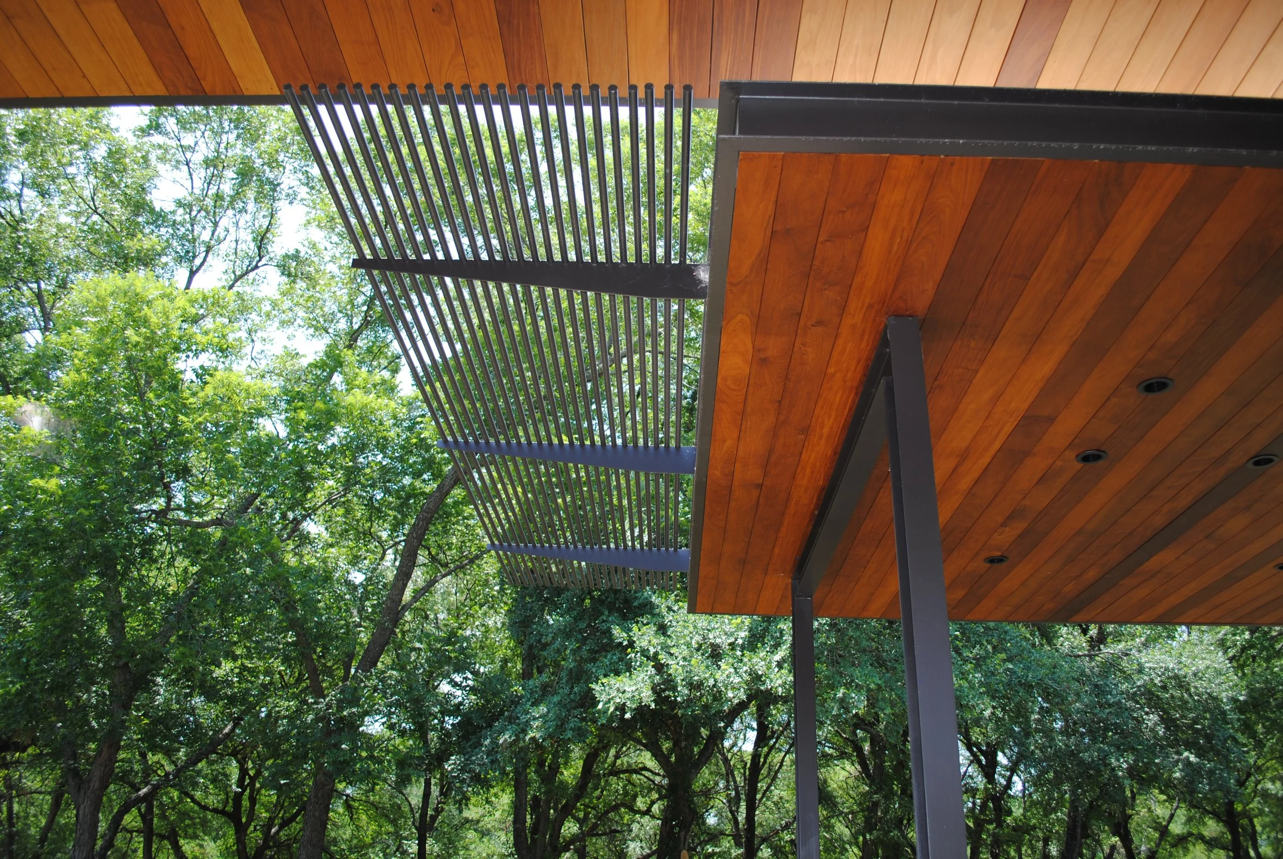 A modern outdoor patio with a wooden ceiling, metal support beams, and a decorative metal railing. Green trees are visible in the background.