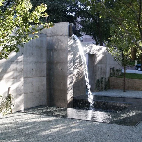 Modern concrete aqueduct-style fountain with water cascading from a bronze spout into a shallow pool, surrounded by trees and outdoor seating.