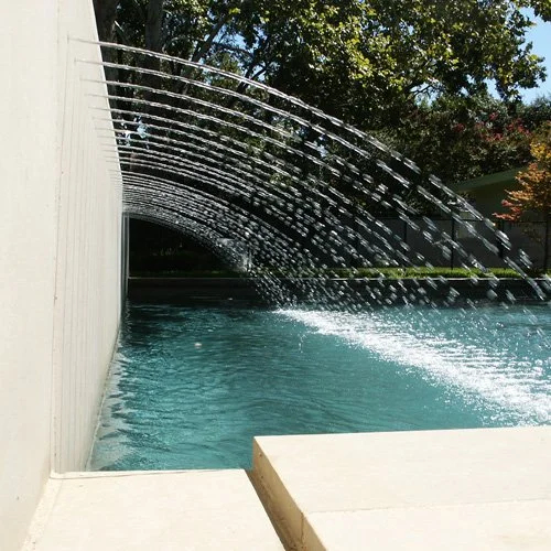 A backyard swimming pool with water arching from spouts along a wall into the pool, with trees and greenery in the background.